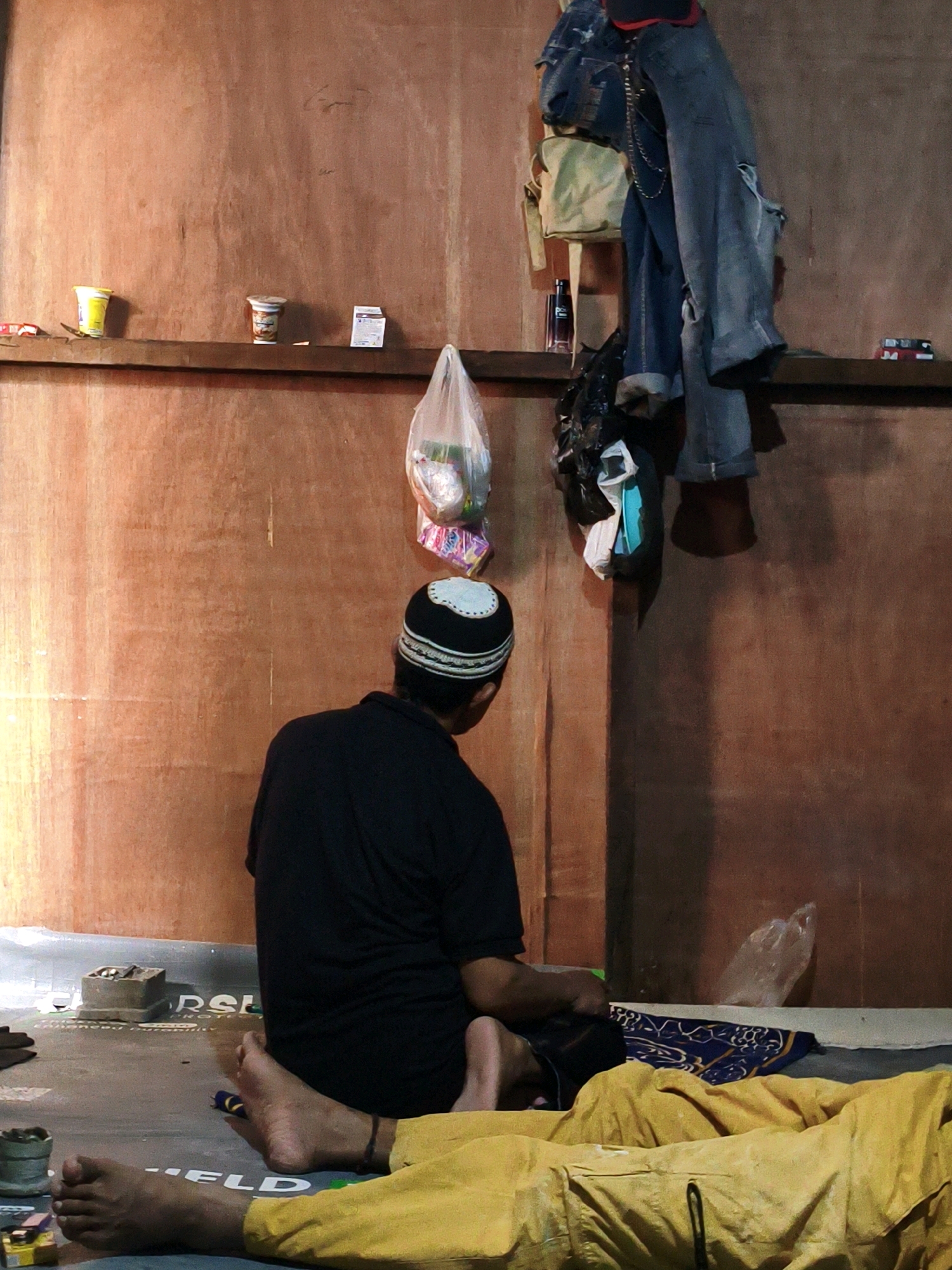 The image depicts an interior scene in Bekasi, Indonesia, likely a modest dwelling or shelter. A person wearing a black t-shirt and a white and black kufi hat is seen from behind, seated on the floor, facing a wooden wall. Their knees are bent, and their hands are resting in their lap, suggesting a moment of rest or prayer.
In the foreground, another person is lying down, covered with a yellow blanket. Their legs are visible, one bare and the other partially covered by the blanket. The floor is covered with a gray material, possibly a tarp or mat, with some text visible, including "RSE" and "ELD".
The wooden wall is sparsely decorated. A narrow shelf runs across the upper portion, holding several items: a yellow cup, a small container with a lid, and a small white packet. Further to the right, a backpack and several pairs of jeans are hanging from what appears to be a hook or nail. A plastic bag containing items is also suspended from the wall.
The lighting is dim, casting shadows on the wall, and giving the scene a somber atmosphere. The overall impression is one of humble living conditions.