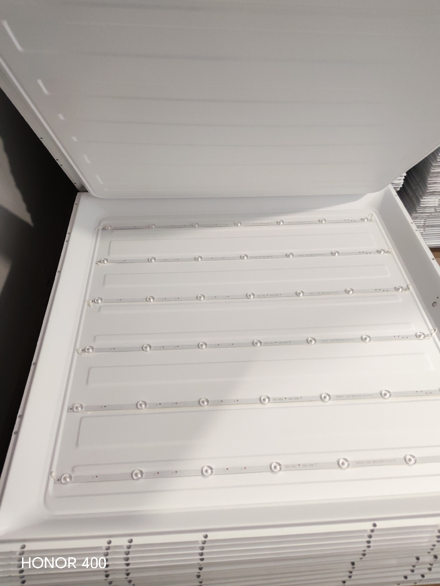 The image shows a close-up, eye-level view of the inside of a large, flat, white LED light fixture. The fixture appears to be rectangular and is oriented horizontally in the frame. It is composed of a white, ribbed surface with several thin LED strips running parallel to each other. Each LED strip has multiple small, circular lenses, presumably covering individual LEDs. The lighting appears to be from an overhead source, casting soft shadows within the grooves of the fixture. The overall impression is one of a new or clean electronic device. The bottom left corner of the image contains the text "HONOR 400" in white, sans-serif font. The background to the left is a dark, out-of-focus area, possibly a wall or shadow. To the right, a stack of similar white objects is visible, also out of focus. The image does not contain any specific geographical or contextual information that would indicate its location in Tashkent, Uzbekistan.