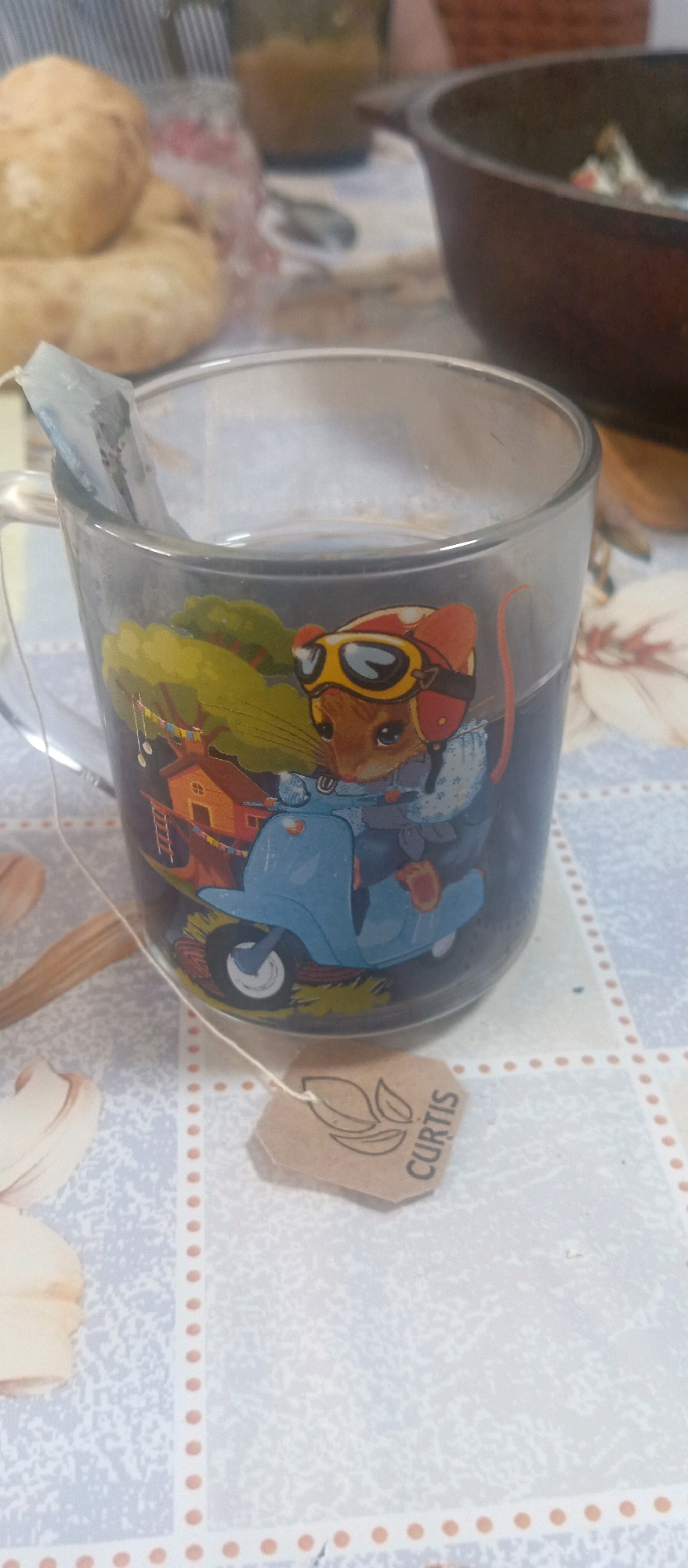 A close-up, eye-level shot captures a clear glass mug filled with tea. The mug is decorated with a graphic of a mouse wearing a helmet and goggles, riding a scooter. A tea bag, with the string hanging over the rim, is steeping in the tea. A small, brown paper tag with "CURTIS" printed on it is attached to the tea bag string and rests on the mug's surface. The mug is placed on a table covered with a light-colored tablecloth featuring a pattern of small, light grey and white shapes. In the background, out of focus, are stacks of light brown bread, a clear container with a greenish-yellow substance, and a dark brown pot. The overall lighting is dim. The photograph was taken in Kunashak, Russia.