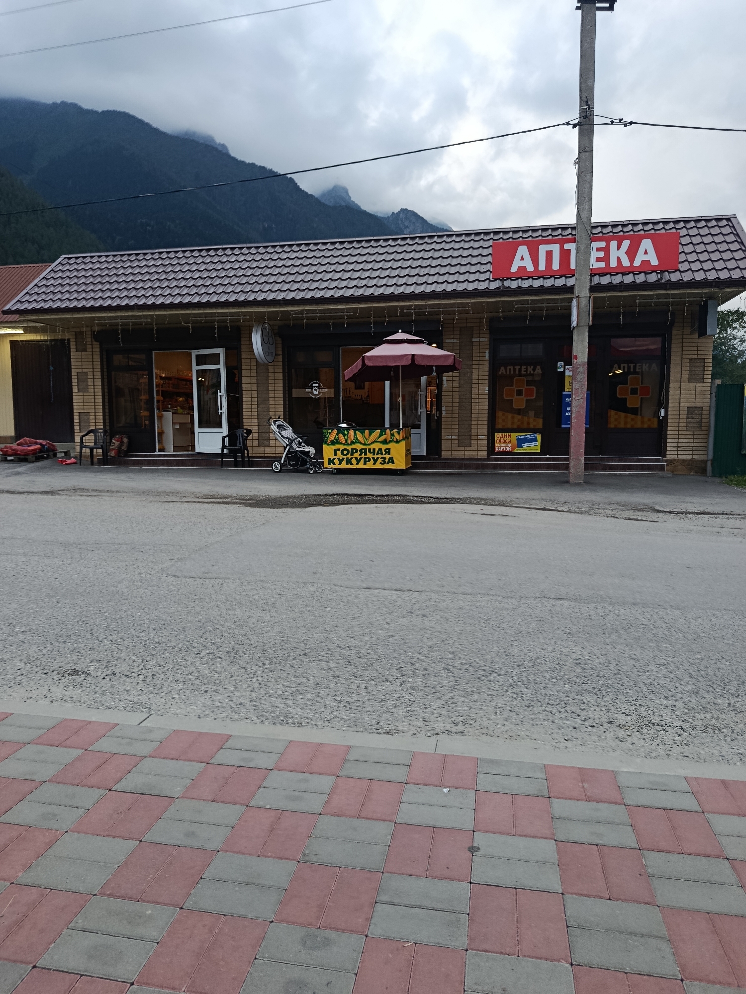 A medium shot captures a single-story building with a brown, tiled roof, set against a backdrop of misty mountains and a cloudy sky. The building's facade is constructed of light brown brick, and two storefronts are visible. The left storefront has a white door and is illuminated from within. The right storefront has a dark brown door with "АПТЕКА" (Apteka, meaning pharmacy) written in white capital letters above it, accompanied by a red cross symbol on the glass. A red awning is extended over a display of corn in front of the left storefront. A baby stroller is positioned to the right of the corn display. A utility pole stands in front of the building, obscuring a portion of the right storefront. In the foreground, a paved walkway of red and grey interlocking bricks leads towards the street, which is made of asphalt. The scene appears to be a street-level view of a commercial area in Teberda, Russia.