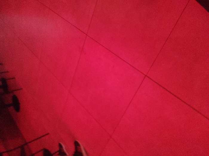 Red-toned image shows a tiled floor, angled slightly, with dark lines separating the tiles. On the left, blurry figures appear, possibly including workout equipment like dumbbells and the feet of a person wearing sneakers. The red hue obscures the details, giving the scene a dark, indistinct ambiance.
