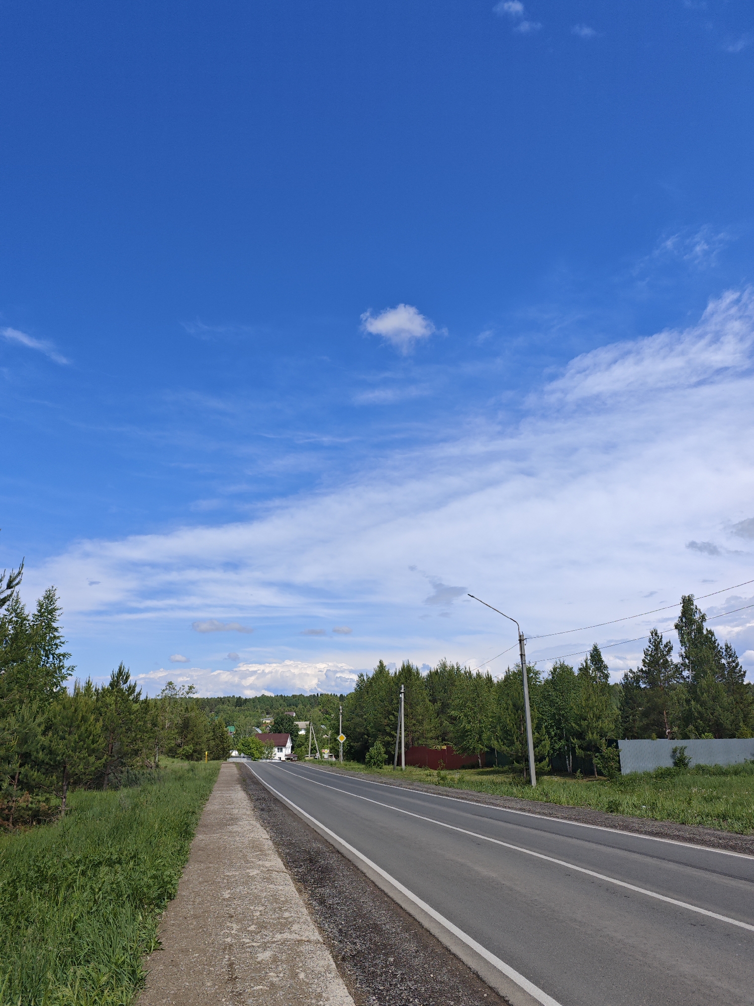 A clear, daytime landscape photograph shows a straight asphalt road running through Марковский, Russia. A concrete sidewalk runs along the left side of the road, bordered by grass. On the other side of the road, trees and fences line the roadside. A power pole stands to the right of the road. In the distance, houses are visible. The sky is predominantly blue with scattered white clouds.