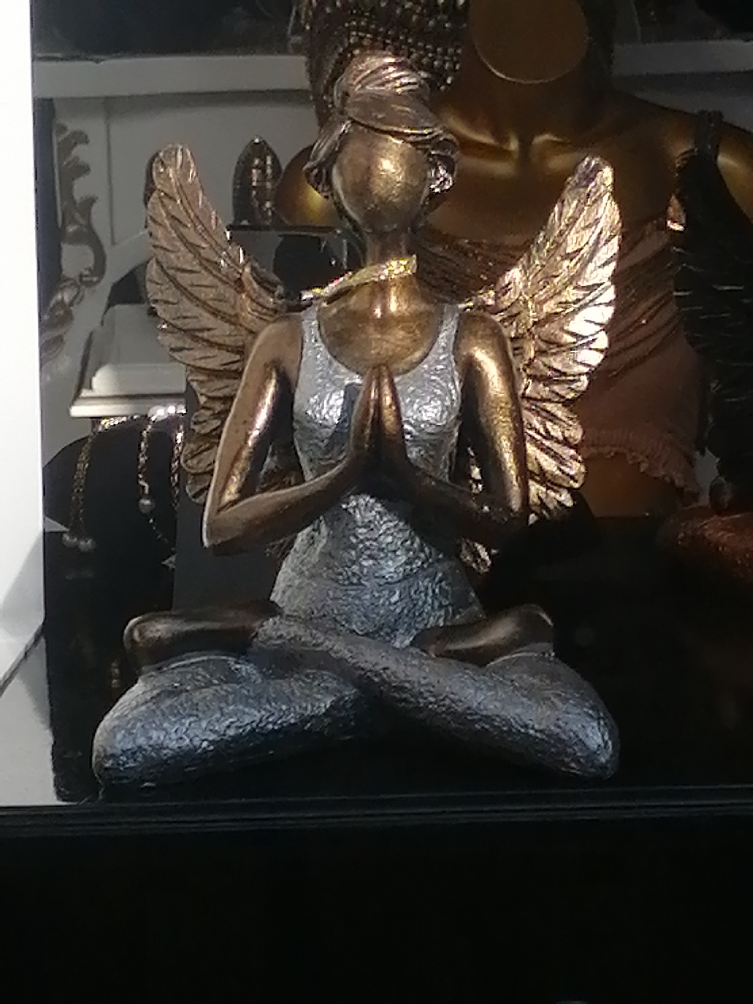 This is a medium shot, slightly low-angle, eye-level photograph of a decorative figurine of a winged woman in a meditative pose. The figurine is made of metallic-finished material, with the body and wings appearing bronze or gold, and the dress a textured silver. The woman is seated with her legs crossed, hands pressed together in front of her chest, and her head slightly bowed. She wears a gold necklace and a metallic head covering. The figurine is positioned in the foreground, with a blurred background that suggests a display or store setting. There are other figurines and decorative items visible in the background, also with metallic finishes. The lighting appears to be artificial, casting subtle reflections on the figurine's surface. The location in the city of Ubud District, Indonesia is not visually evident in the photograph itself.