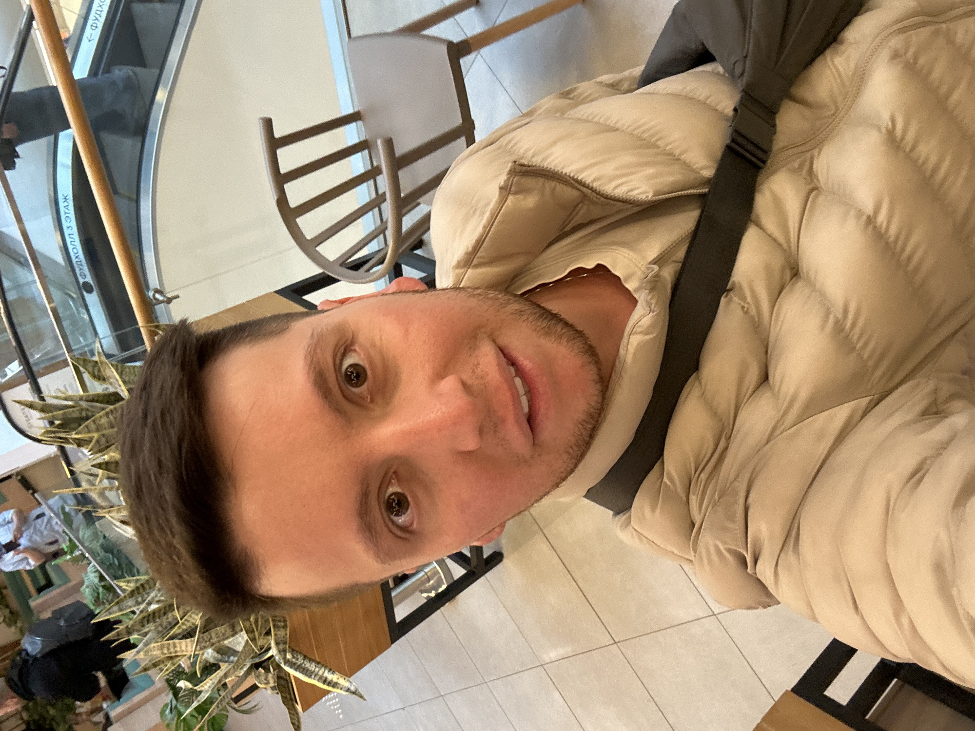 The image shows a first-person perspective selfie of a man with brown hair and brown eyes, wearing a beige puffer jacket with a black strap across his chest. He is indoors, likely in a shopping mall, as an escalator is visible in the background with signage in Cyrillic script. There are plants to the left of the man. The floor is tiled. The man is positioned near a chair with wooden slats. Based on the Cyrillic script visible on the escalator, the location is likely in Moscow, Russia.