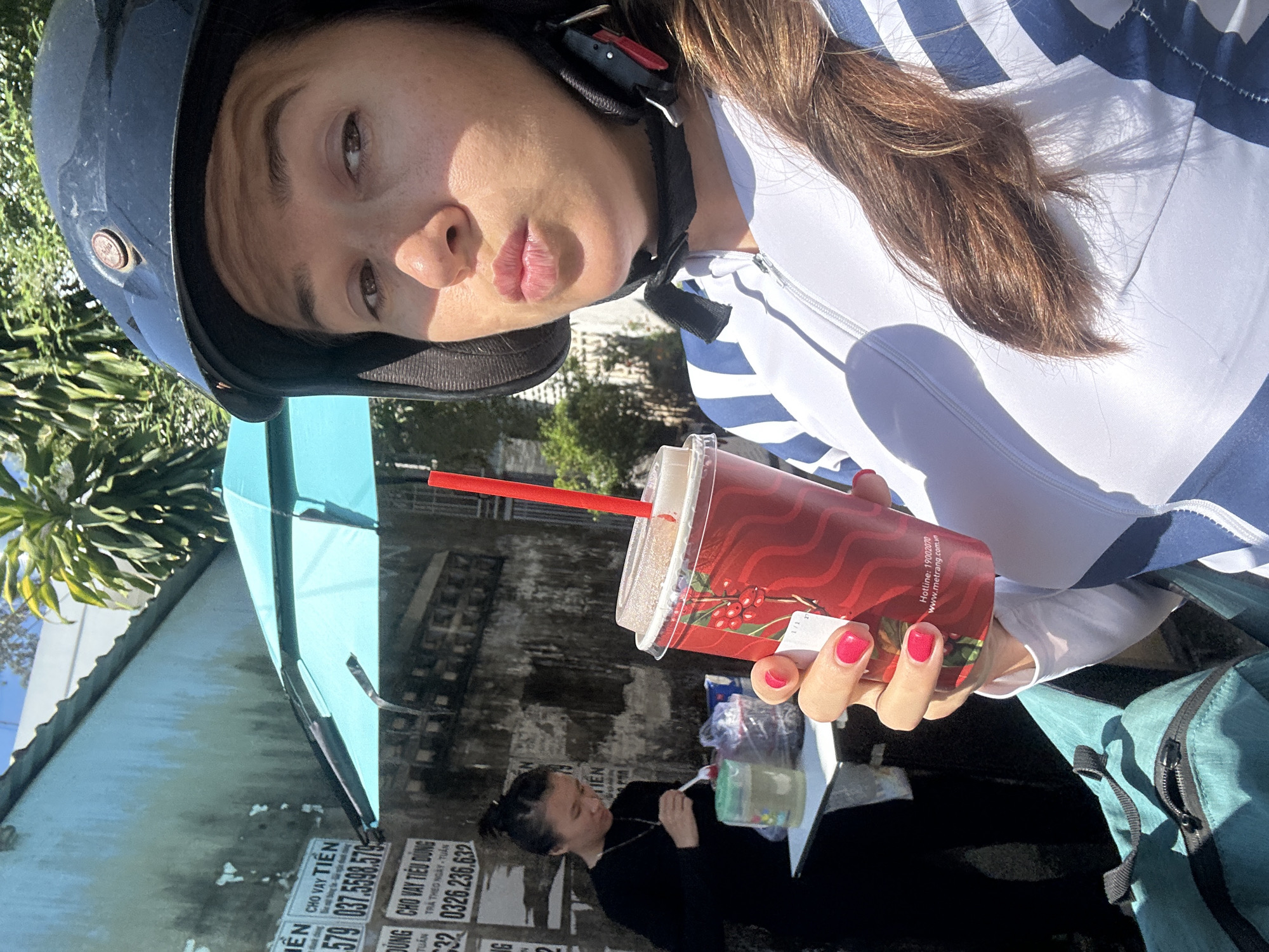 The image shows a woman wearing a helmet and a white-and-blue striped shirt. She is holding a red cup with a red straw. Her nails are painted red. Behind her, there is a street scene with a vendor eating at a table. There are signs with Vietnamese writing on a wall. The OCR indicates the signs advertise loan services and include contact information. The cup has writing in Vietnamese, including "Hotline: 19002070" and "www.metrang.com.vn." Based on the signs and text, the location is likely in the city of Nha Trang. It is likely Bac Nha Trang Ward, Vietnam.
