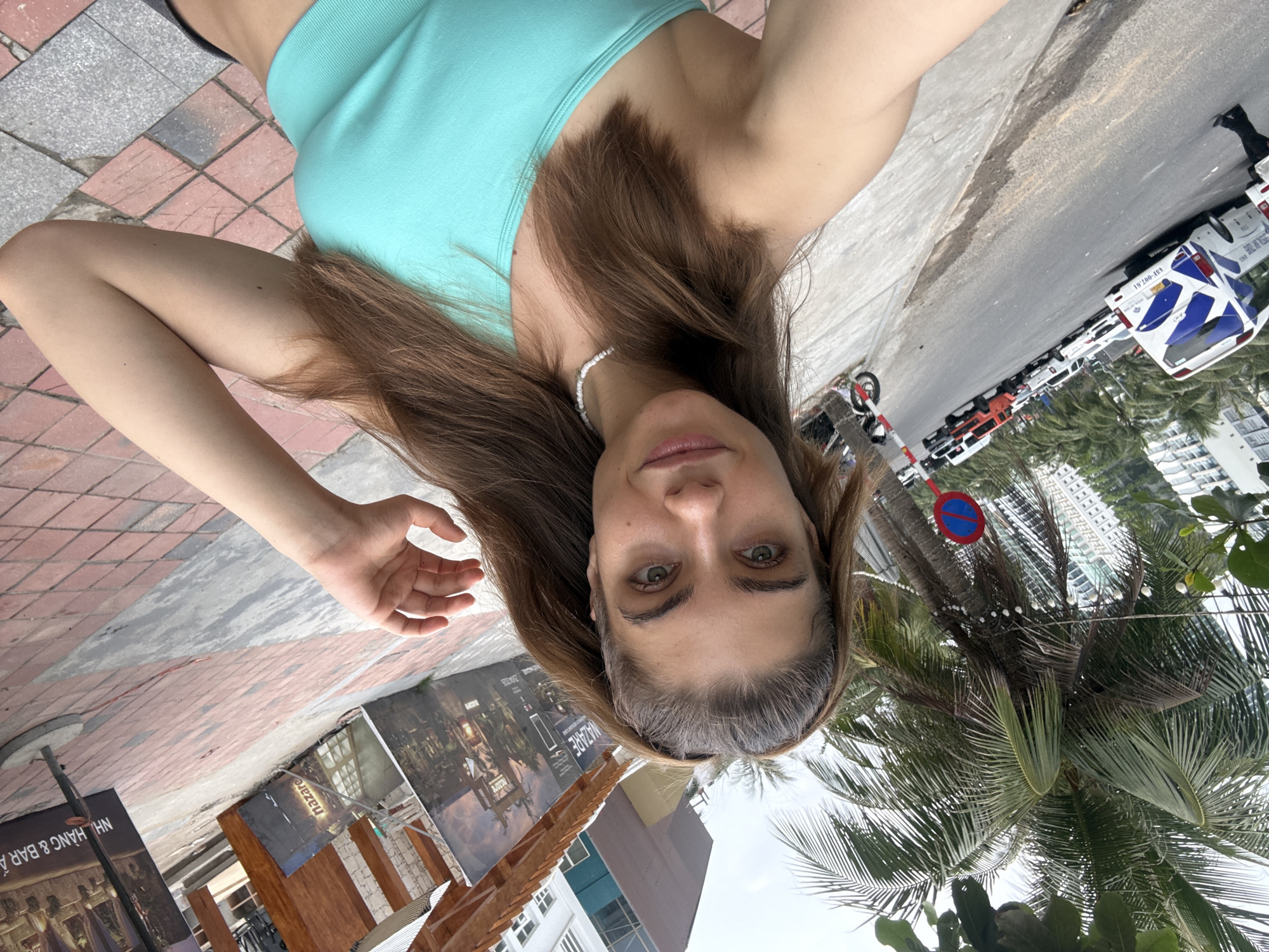 A selfie of a woman in Đà Nẵng, Vietnam. The woman is fair-skinned with brown hair and is wearing a light blue top. In the background is a street scene with parked cars, including a police vehicle, palm trees, and buildings. There are signs indicating local businesses and streets. The photo appears to be taken on a sidewalk or pedestrian area.