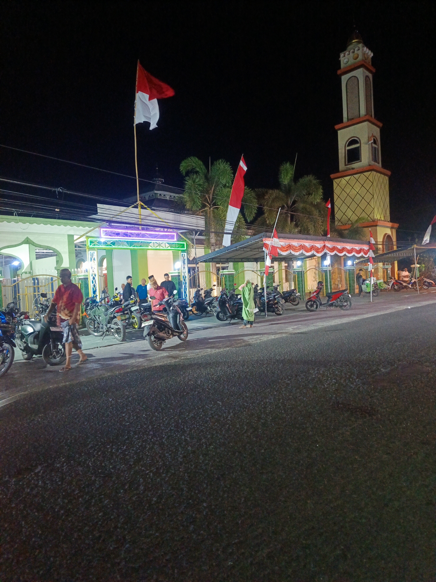 The image depicts a nighttime scene in Gorontalo, Indonesia, likely outside a mosque, given the presence of a minaret and the architectural style of the building in the background. The scene is illuminated by artificial lights, casting a warm glow on the surroundings. In the foreground, a paved road is visible, with several parked motorcycles lined up along the roadside. A few people are present, some of whom appear to be standing near the motorcycles, possibly engaged in conversation or waiting.
Further back, the mosque building is partially visible, featuring a light green and white color scheme. The entrance to the mosque is adorned with colorful neon lights, creating a festive atmosphere. Indonesian flags, red and white, are prominently displayed on flagpoles, suggesting a national celebration or event. The minaret of the mosque stands tall in the background, a distinctive architectural element of Islamic places of worship. Palm trees are scattered throughout the scene, adding a tropical touch to the environment. The overall impression is one of a community gathering at night, possibly for a religious or cultural event.
