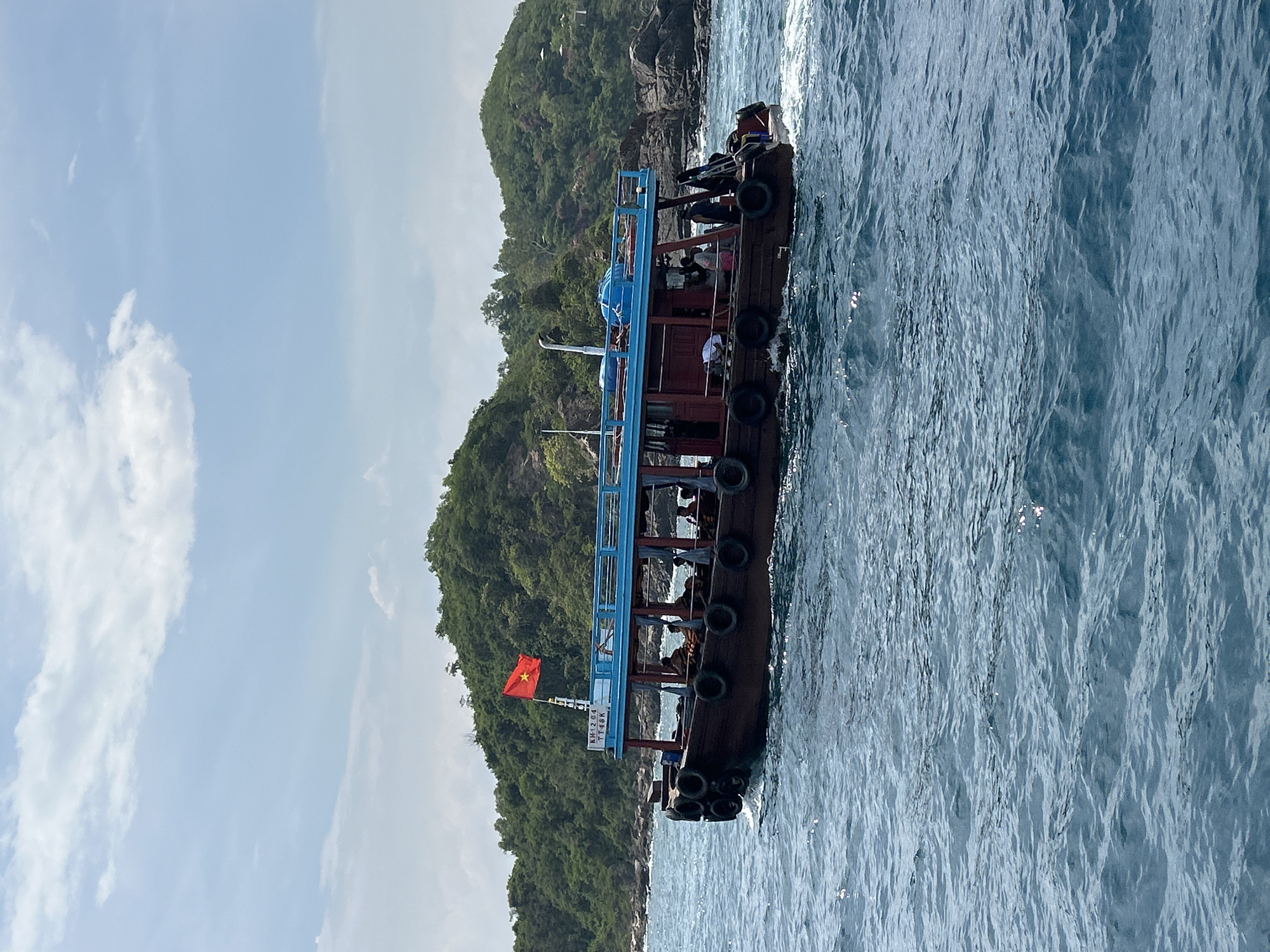 A wooden boat with a blue roof sails on the sea near Nha Trang, Vietnam. The boat carries passengers and flies the Vietnamese flag. Lush, green hills rise from the rocky shoreline in the background under a partly cloudy sky. The water is a deep blue with gentle waves.