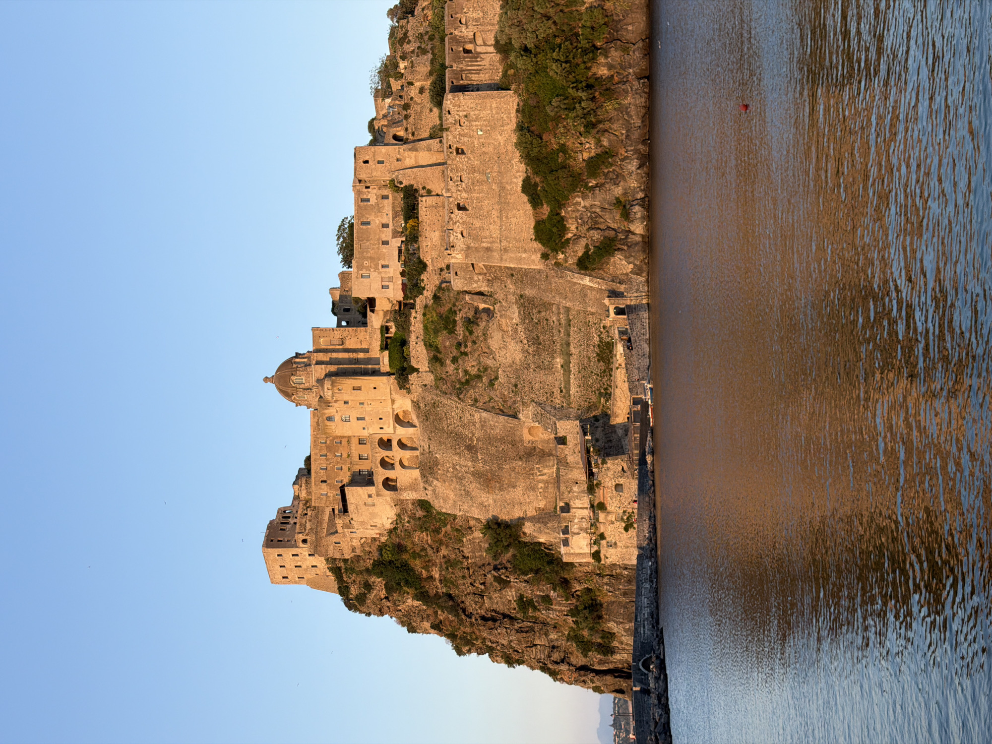 The image depicts the Aragonese Castle, a medieval fortress located on a volcanic rocky islet connected to Ischia, Italy, by a causeway. The castle complex consists of interconnected structures built on the island's rugged terrain. The buildings are primarily constructed from stone, displaying various architectural styles accumulated over centuries. The islet's steep cliffs are covered in vegetation, including trees and shrubs. The surrounding water is calm, reflecting sunlight and the structures of the castle, indicating a time of day with good lighting conditions. The sky is clear and blue, providing a backdrop to the castle.