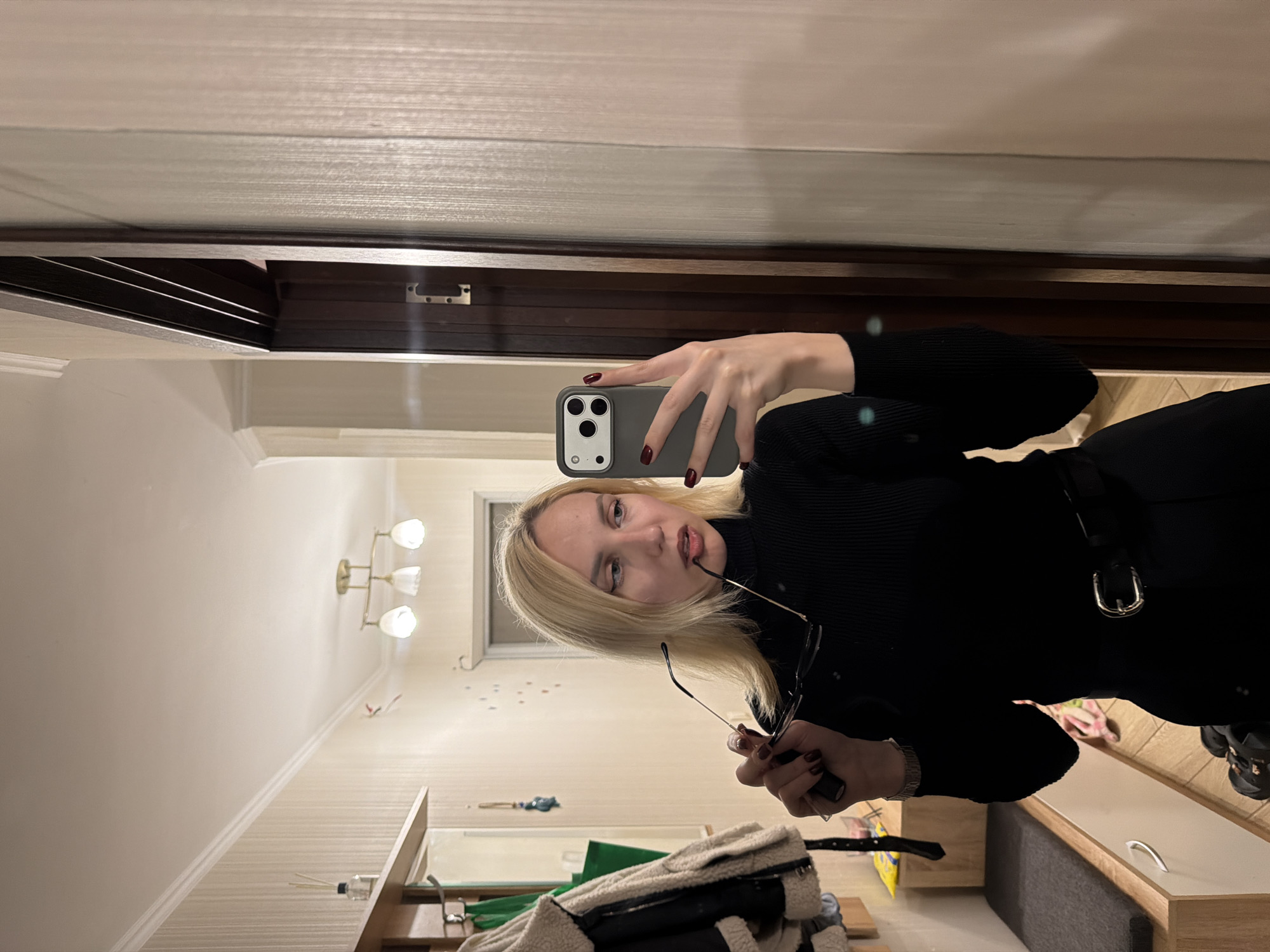 A young woman with blonde hair is taking a selfie in a hallway. She is wearing a black turtleneck and black pants with a belt. She is holding a pair of glasses in her right hand and her phone in her left hand. The hallway appears to be in an apartment building, with a coat rack, a bench, and a mirror visible in the background. The lighting is dim and artificial. There are no visible indications of the city or country.