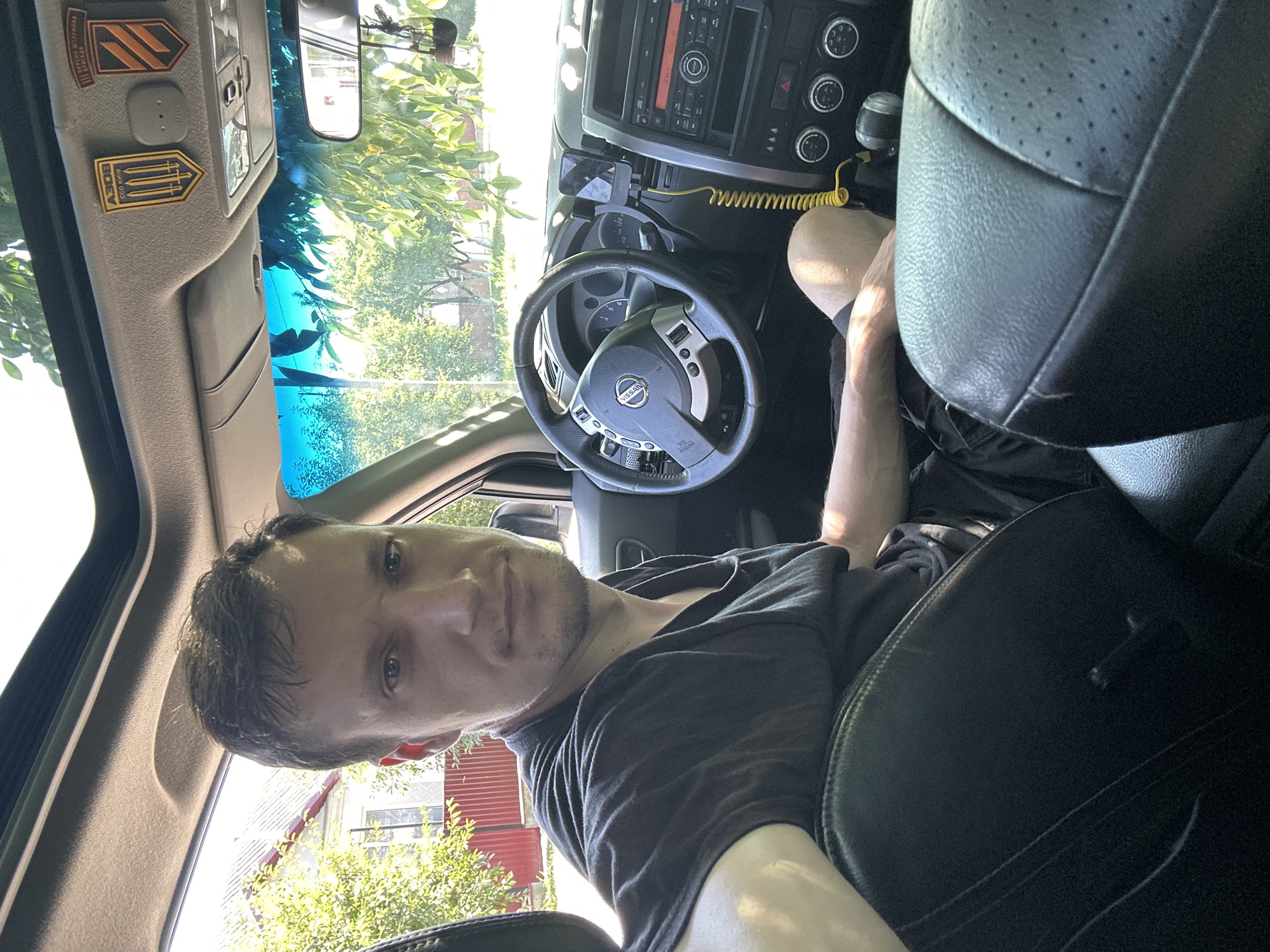 A man is sitting in the driver's seat of a car. He is wearing a black t-shirt and black shorts. The car is parked on a street in Shatava, Ukraine. The street is lined with trees and houses. The man is looking at the camera. The car has a sunroof and a rearview mirror. There are two patches on the car's visor, one with the letters "АЗОВ" and a symbol of three swords, and another with orange and black stripes. The man's left arm is resting on the driver's seat. The car's dashboard is visible, with a Nissan steering wheel, a radio, and air conditioning controls.