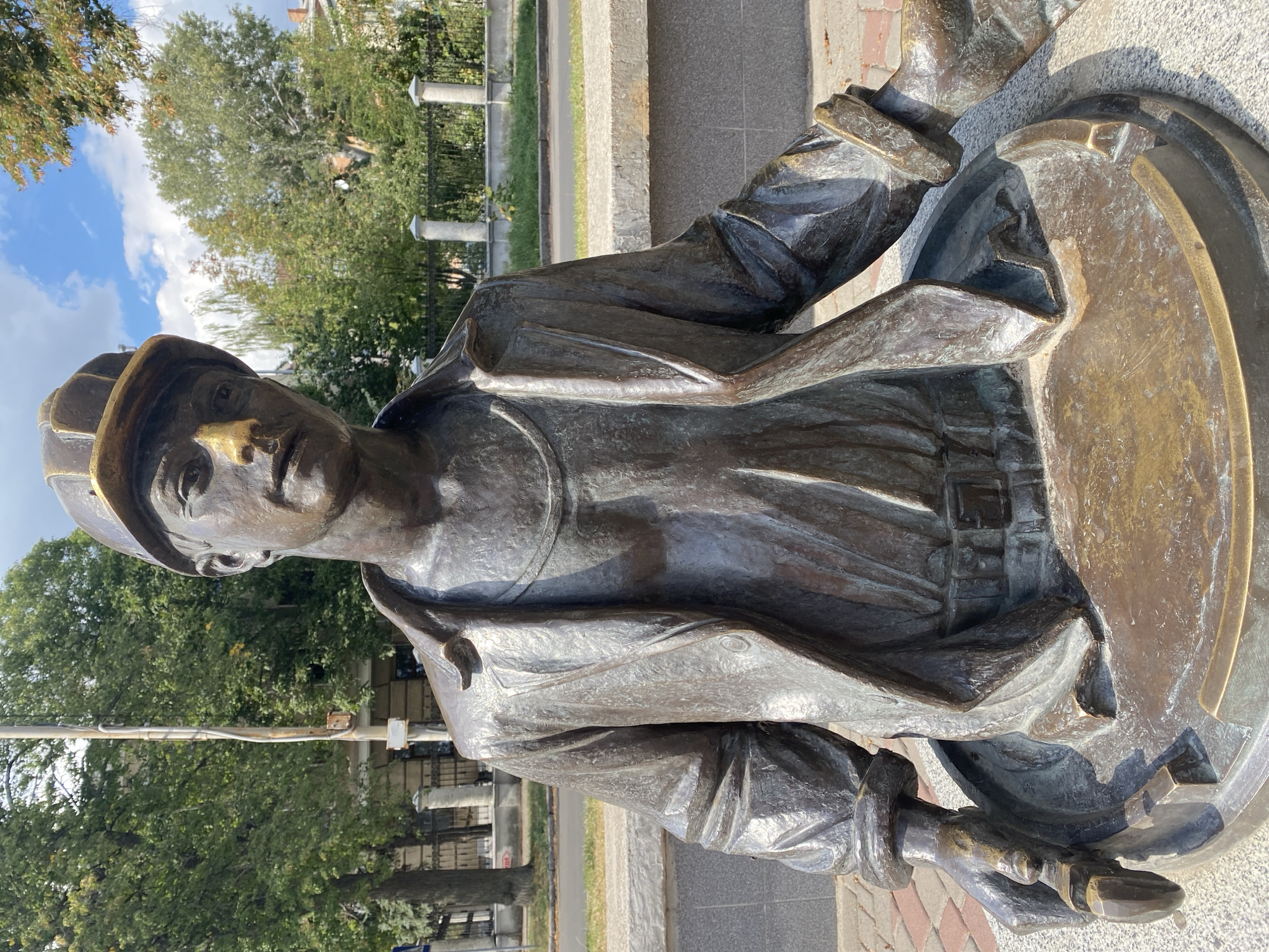 A bronze sculpture of a male construction worker is positioned on a circular base, appearing to emerge from a manhole cover. He wears a hard hat, a jacket, and a shirt with a chain visible around his neck. His right hand holds a pipe wrench, and his left hand is positioned as if resting on the edge of the manhole. The sculpture is located in the city of Poltava, Ukraine, near a street with a sidewalk and a metal fence with ornate pillars in the background. Trees and a building are visible behind the fence. The sculpture is situated on a paved area with a brick pattern. The sky above is blue with scattered clouds.