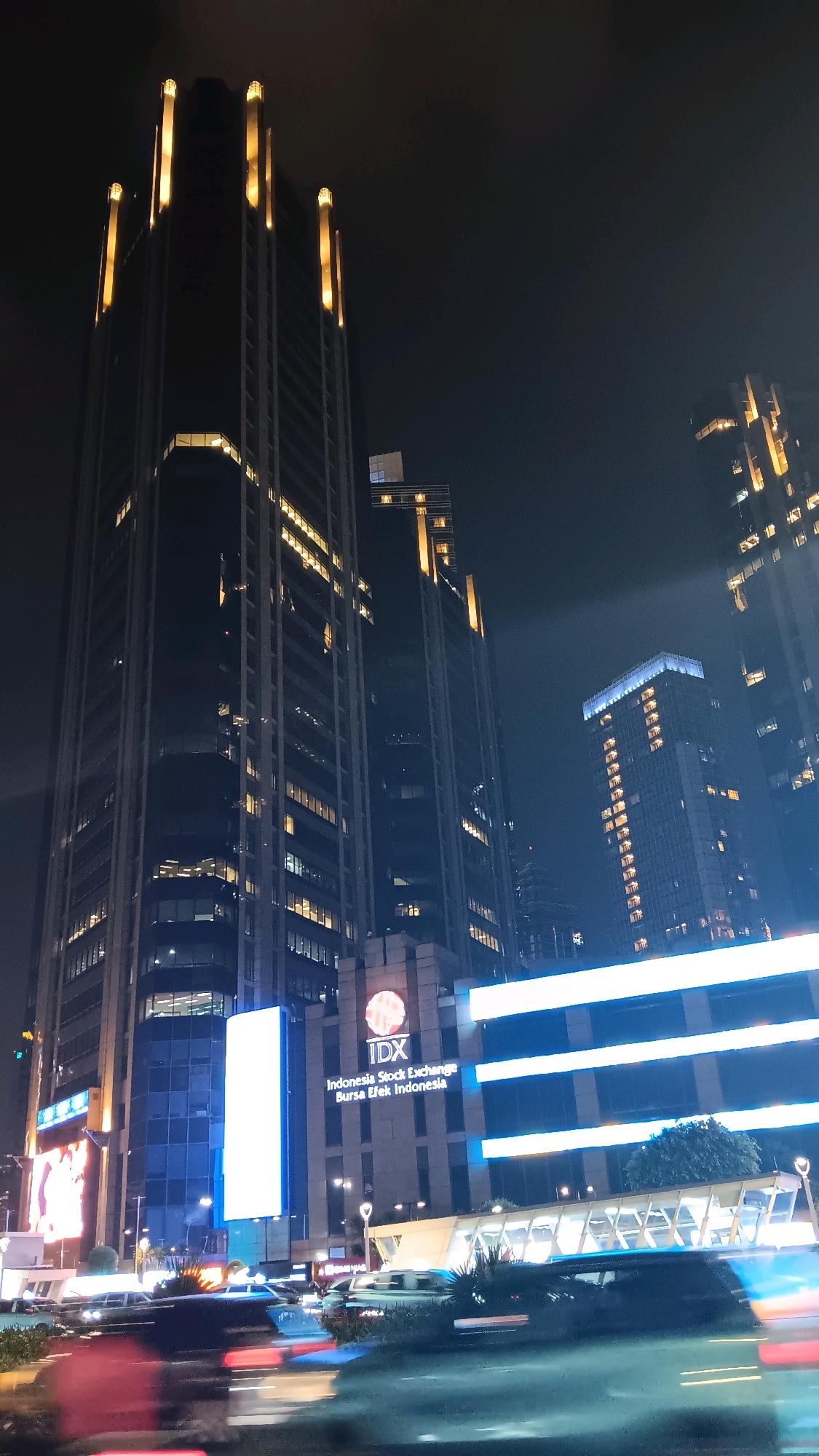A nighttime, low-angle, vertical shot captures the Indonesia Stock Exchange (IDX) building in Jakarta, Indonesia. The skyscraper, illuminated by yellow lights at its apex and with numerous windows lit from within, dominates the frame. To its right, another skyscraper also features illuminated windows. In the foreground, a street scene shows blurred car headlights and taillights, indicating motion. The lower portion of the IDX building is clearly visible, displaying the building's logo and name in white text against a facade. Blue and white lights outline a structure in front of the building. The sky is dark and hazy, suggesting a humid night.