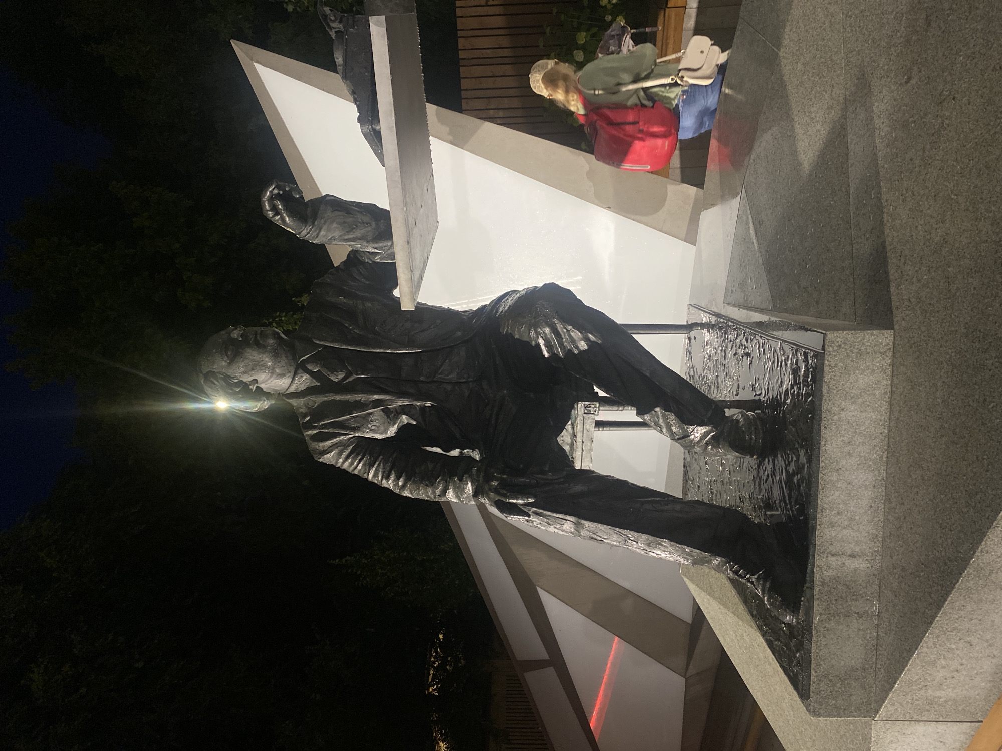 A bronze statue of a man in a suit is seated on a stool, with his left hand resting on a table. The statue is in a public space at night, illuminated by artificial lights that create glare. A young woman with a red backpack and a beige purse stands to the right of the statue, looking away from the camera. The statue is located in Novokuybyshevsk, Russia. The base of the statue is a granite platform, and there is water on the ground around the statue's feet. A white angled structure forms the background behind the statue.
