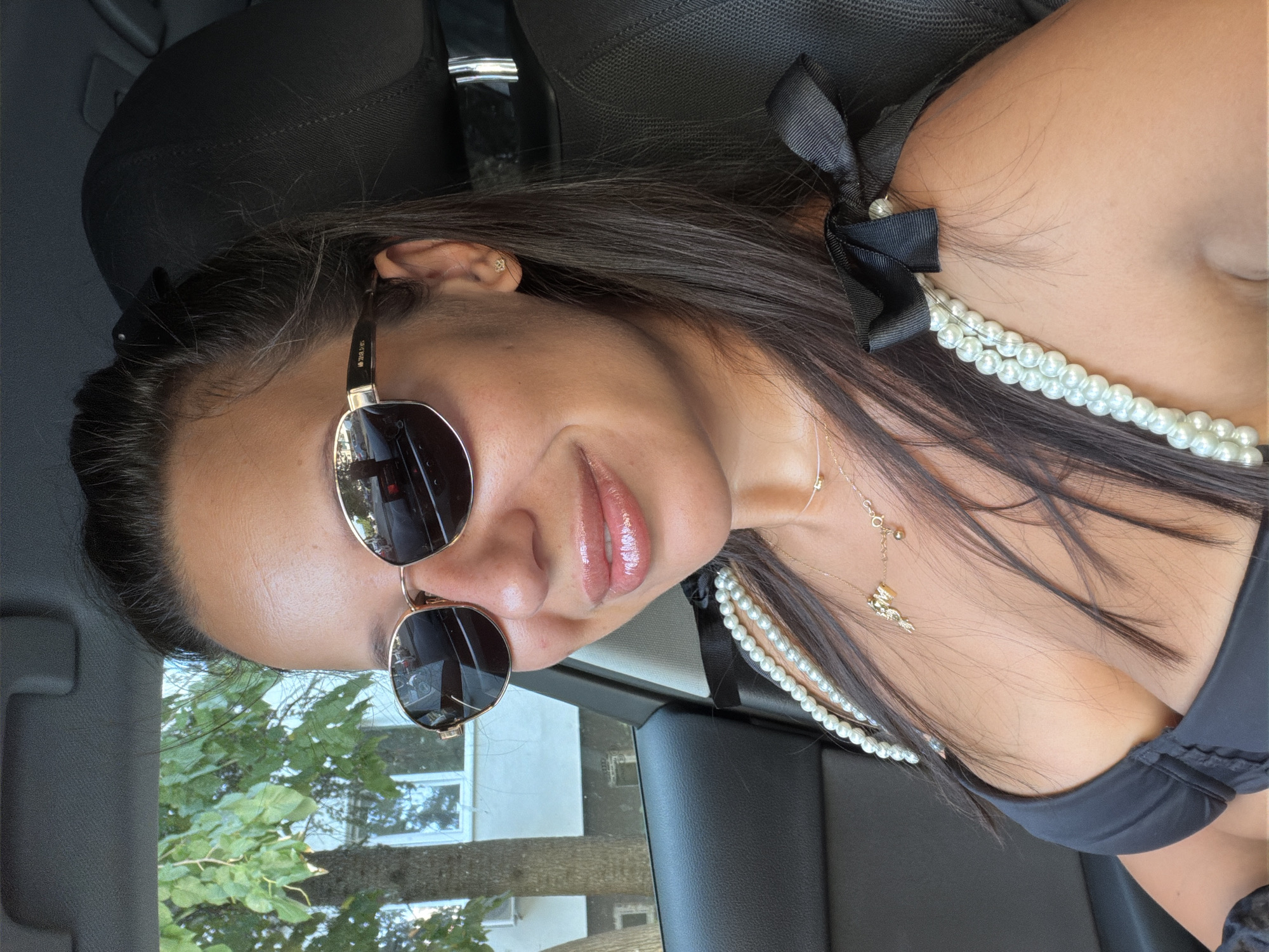 A woman is sitting in the driver's seat of a car. She is wearing sunglasses, a black top, and a pearl necklace. Her hair is dark brown and pulled back. She is smiling and looking at the camera. The car is parked on a street in Burgas, Bulgaria. A tree and a building are visible in the background.