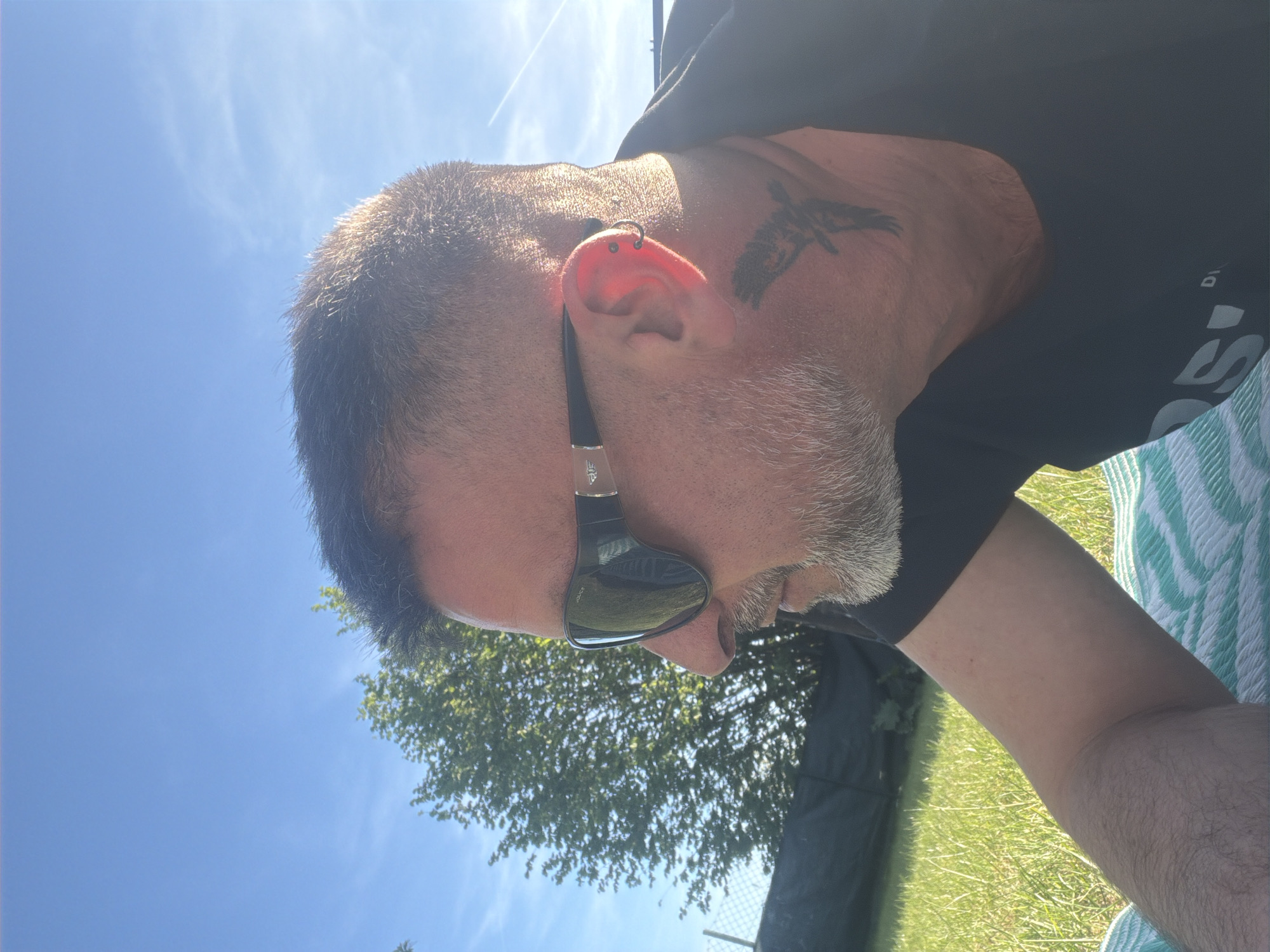 A man with grey hair, a grey beard, and sunglasses is positioned in an outdoor setting, seemingly in Junglinster, Luxembourg. He is wearing a black t-shirt. He has an ear piercing and a tattoo on the side of his face. The background includes a blue sky, trees, and grass. The man appears to be reclining on a green and white striped blanket.
