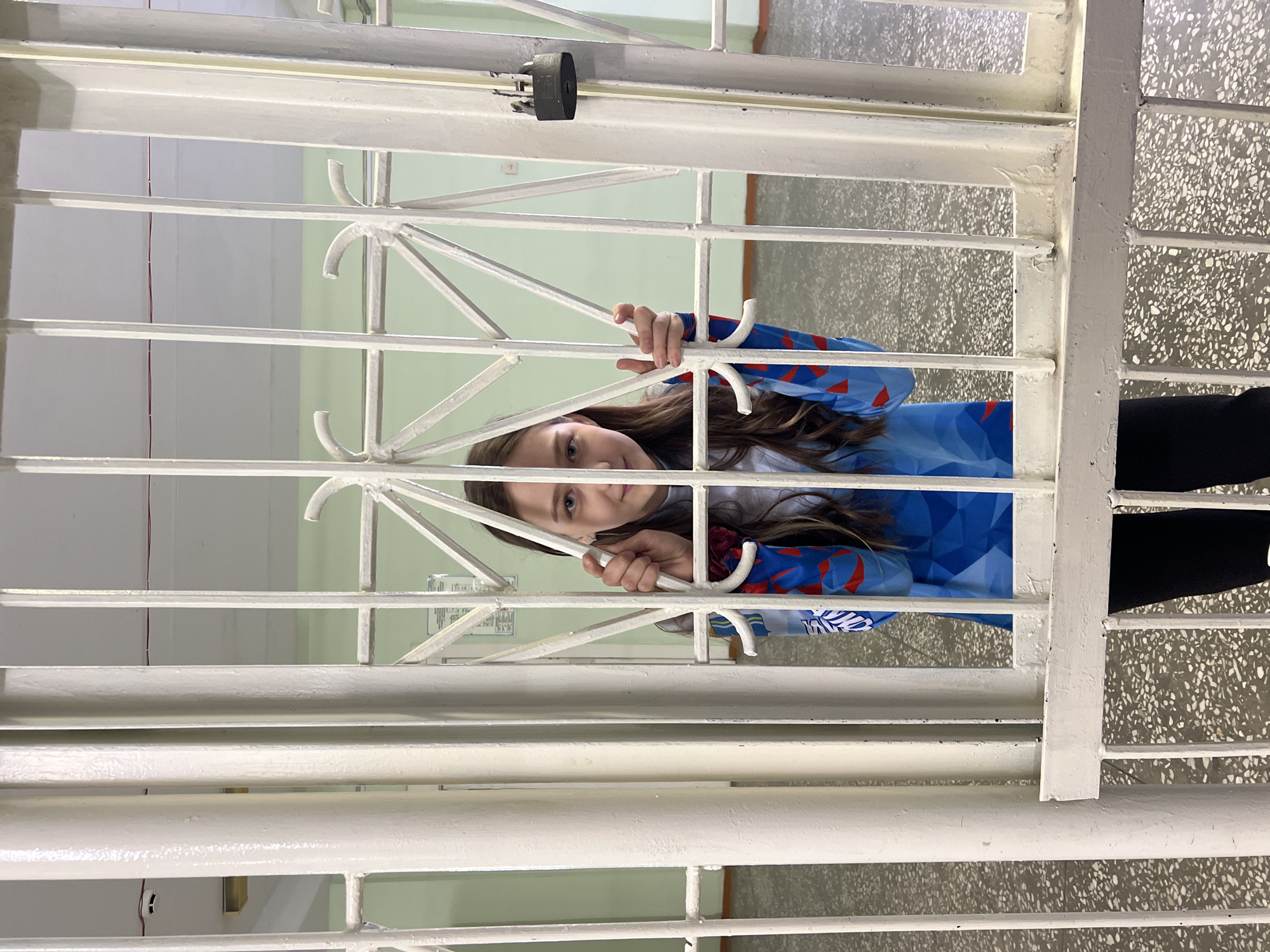 A girl is standing behind a set of white iron bars. She is holding onto the bars with both hands, her face visible between them. She is wearing a blue and red patterned shirt. Part of a black garment is visible on the right side of the frame. The background consists of a room with walls painted in muted tones of white and green, and a floor with a speckled pattern. The location is inferred to be Berezniki, Russia.