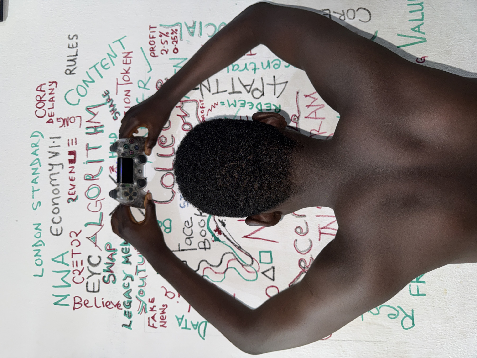 A top-down shot shows a dark-skinned person holding a video game controller. The background is a white surface covered in handwritten words and symbols in red and green ink. The words include "London Standard," "NWA Economy," "Algorithm," "Fake News," "Content," "Profit," "Facebook," and "Ram." The person's head is in the center of the frame, with their arms extending towards the controller. The scene is possibly located in the city of Ado, Nigeria.