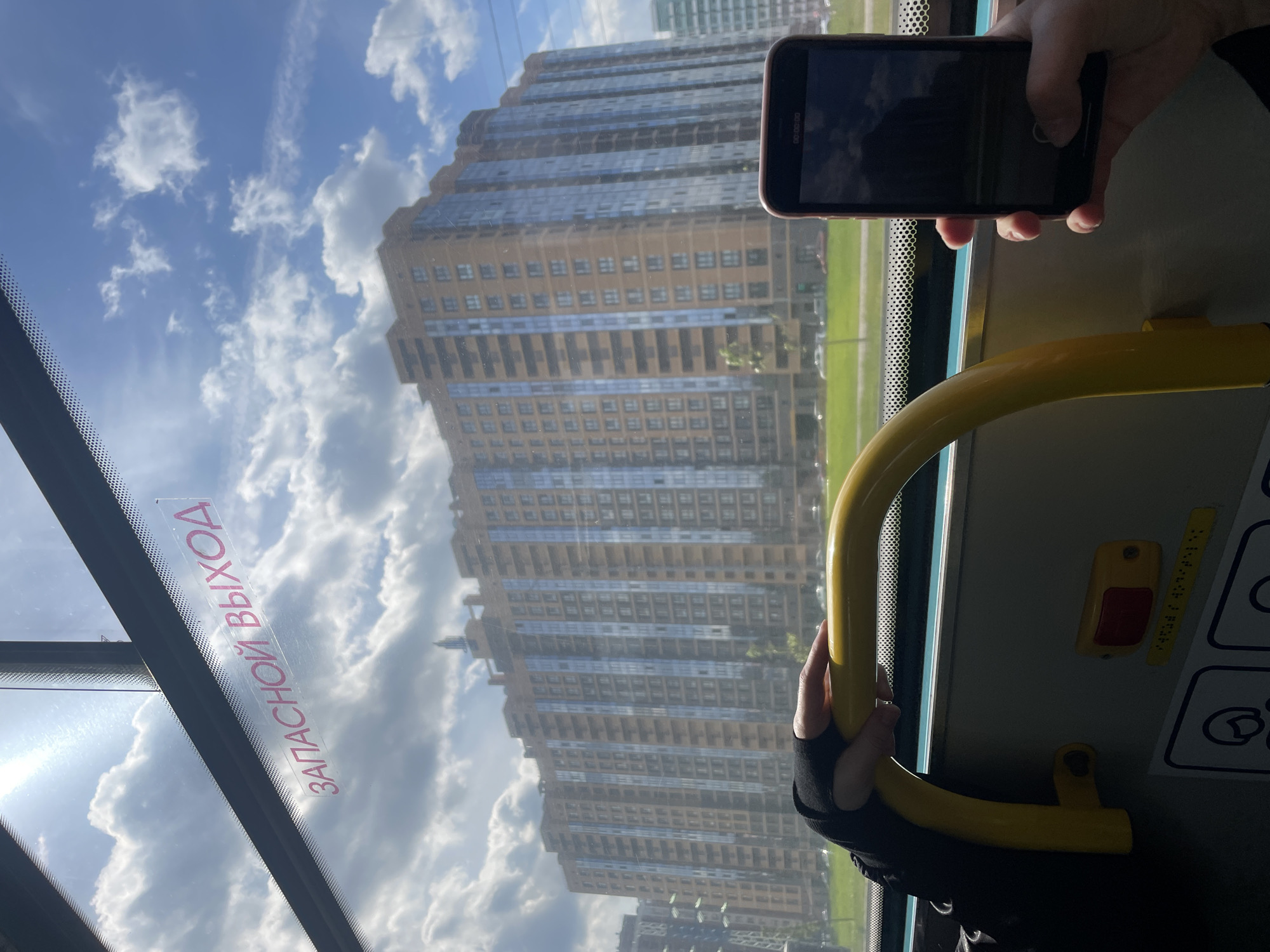 The image captures a bright daytime scene from inside a public transport vehicle, likely a bus or trolleybus, looking out through a window. The perspective is rotated 90 degrees clockwise, showing a tall residential building with many windows and balconies as the prominent exterior feature. The building's facade alternates between sections of beige or light brown cladding and reflective glass panels, rising against a blue sky dotted with scattered white clouds. A thin strip of green grass or land is visible at the base of the building, suggesting a roadside or park area.
Inside the vehicle, a person's left hand, wearing a black fingerless glove, is holding onto a curved yellow grab handle. To the right, another person's hand is holding a black or dark red smartphone. The phone screen is dark but reflects the sky and clouds, and faint digital text "00:00:00" and a circular button suggest a video recording interface. Below the phone, a small red button and other symbols are visible on the vehicle's interior wall.
The window pane features red text, "ЗАПАСНОЙ ВЫХОД", which translates from Russian as "EMERGENCY EXIT", indicating the location is in Russia, specifically Saint Petersburg as per context. The sun is shining brightly, casting reflections and highlighting the clouds. The scene conveys a sense of urban travel on a clear, pleasant day in Saint Petersburg.