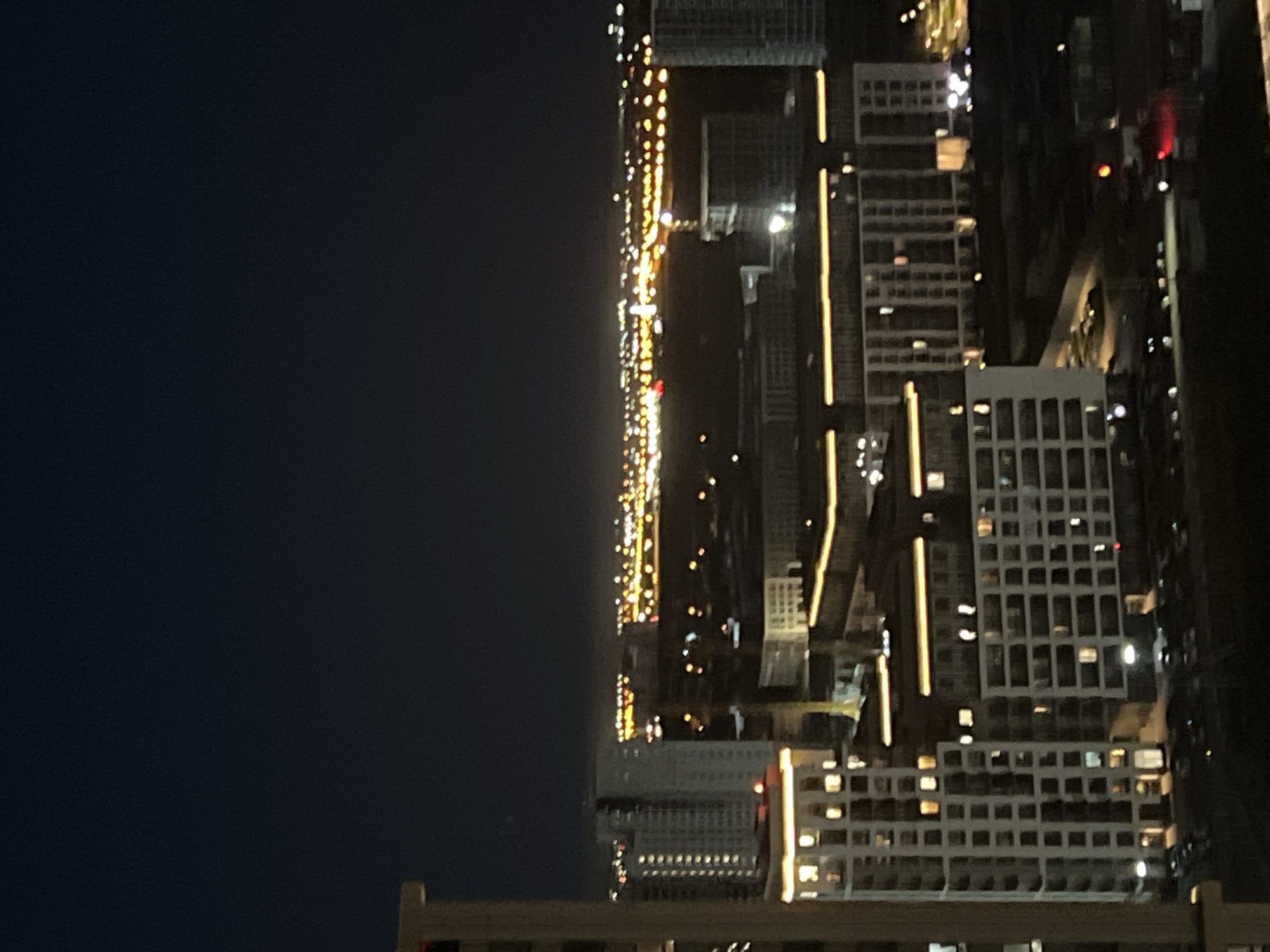 The image shows a night view of a modern urban landscape in Yekaterinburg, Russia. The foreground features a light-colored, multi-story building with numerous windows illuminated from within, suggesting occupancy. To its right, another building with a similar grid-like facade and lit windows is visible. In the background, a sprawling cityscape is illuminated by a multitude of lights, creating a warm glow against the dark night sky. Distant buildings and possibly streetlights create a layered effect, stretching towards the horizon. The overall scene conveys a sense of urban development and activity during nighttime hours.