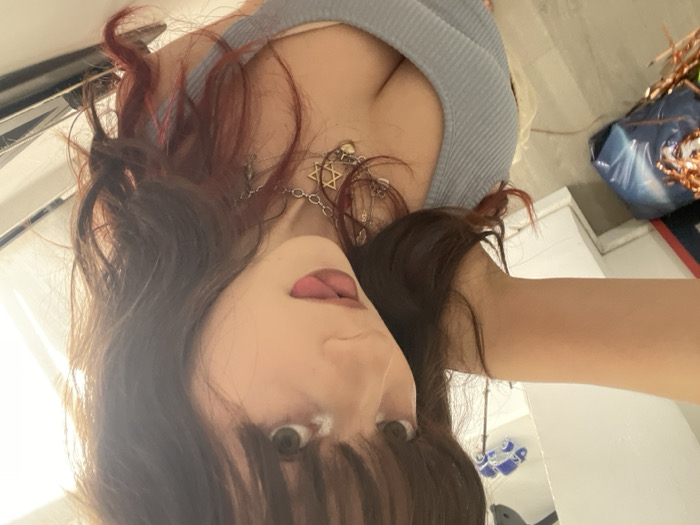 A close-up, selfie-style image depicts a single young female subject indoors, in what is stated to be Raanana, Israel. The subject has dark, shoulder-length hair with bangs and reddish highlights, light-colored eyes with visible eye makeup, and is seen extending her tongue. She wears a light blue ribbed tank top, showing cleavage, and multiple necklaces, one of which features a prominent Star of David pendant. The background is out of focus, revealing a white wall, a light-colored floor, and a bright light source on the upper right. Objects partially visible in the background include a blue-wrapped item with copper-colored material on the lower left floor, and a small blue object resembling a toy in the upper left, against a white vertical surface.