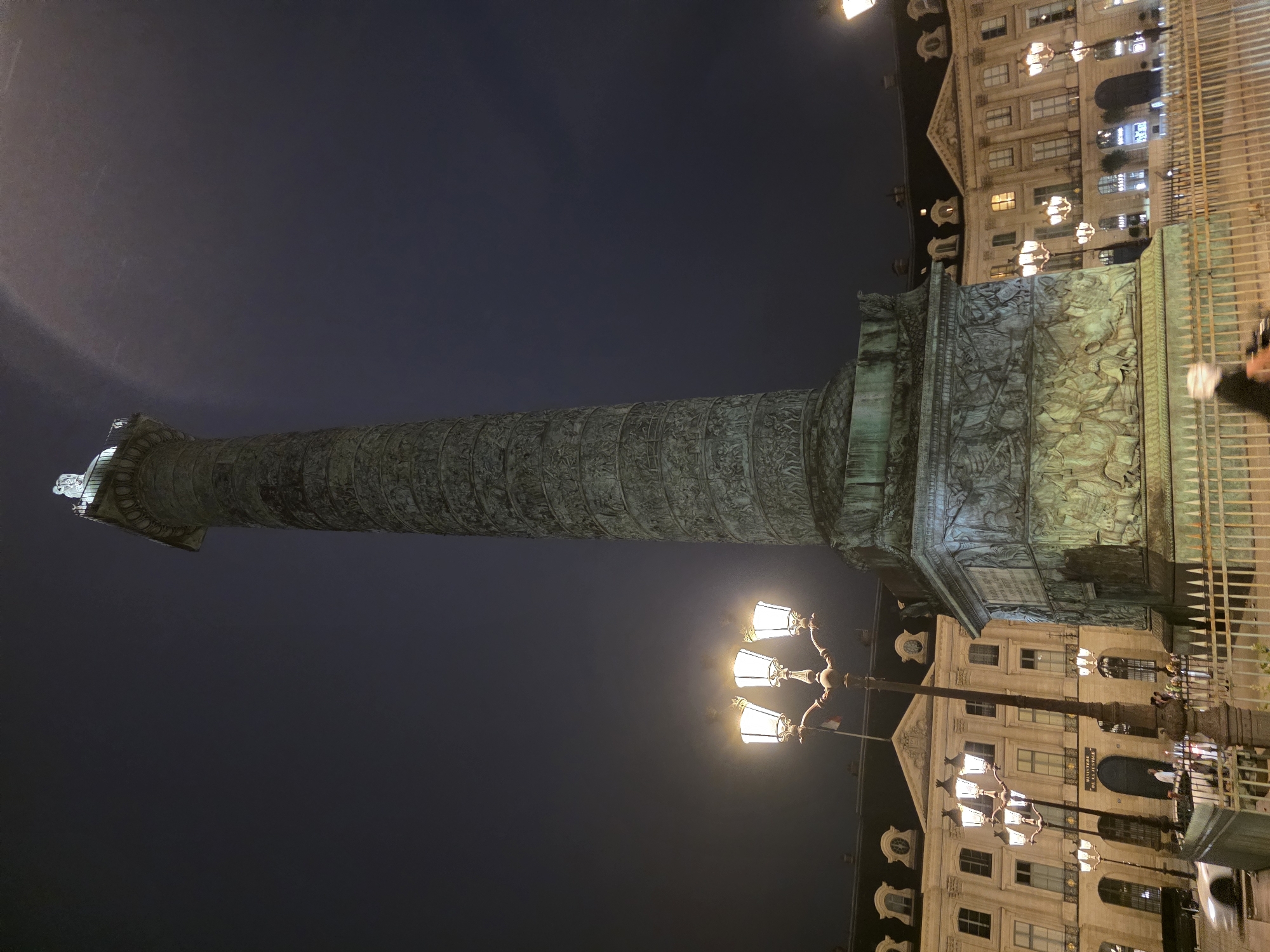 A nighttime, medium-angle shot captures the Vendôme Column in Paris, France. The column, a tall, ornate structure covered in bas-relief carvings depicting Napoleonic victories, dominates the right side of the frame. Atop the column, a statue, likely of Victory, is visible. To the left of the column, a decorative lamppost with three globes casts a warm light on the scene. Behind the lamppost and column, neoclassical buildings with illuminated windows and mansard roofs line the Place Vendôme. A white car is blurred in motion on the street in the lower left. A metal fence encloses the base of the monument. The sky is dark and overcast, with some smudges of light suggesting atmospheric conditions or light pollution. The overall impression is one of grandeur and historical significance illuminated against a dark sky.