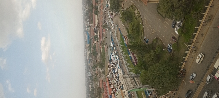 This high-angle, panoramic daytime view captures a bustling urban scene in Nairobi, Kenya. The image, rotated 90 degrees clockwise, depicts a wide city thoroughfare and surrounding cityscape under a partly cloudy to hazy sky.
In the upper portion of the correctly oriented frame, a pale blue sky with scattered white clouds dominates, transitioning to a lighter, hazier tone towards the distant horizon. Below this, a dense urban environment unfolds. A prominent, multi-lane road stretches through the middle and foreground of the scene, populated by numerous vehicles including cars and buses, indicating active traffic flow. A landscaped median with green grass and trees runs down the center of this main road.
To the sides of the main road, more green spaces with mature trees are visible, alongside smaller roads and parking areas filled with parked cars. A dense collection of buildings of various heights and architectural styles extends into the background, hinting at a large and developed city. Small, indistinct figures of people can be observed on sidewalks and near vehicles, though their details are not discernible due to the distance.
The scene conveys general urban activity during daylight hours, possibly mid-morning or afternoon, given the bright illumination and soft shadows. The overall impression is one of a busy, expansive city. No discernible text is visible on any signs, vehicles, or buildings within the image.