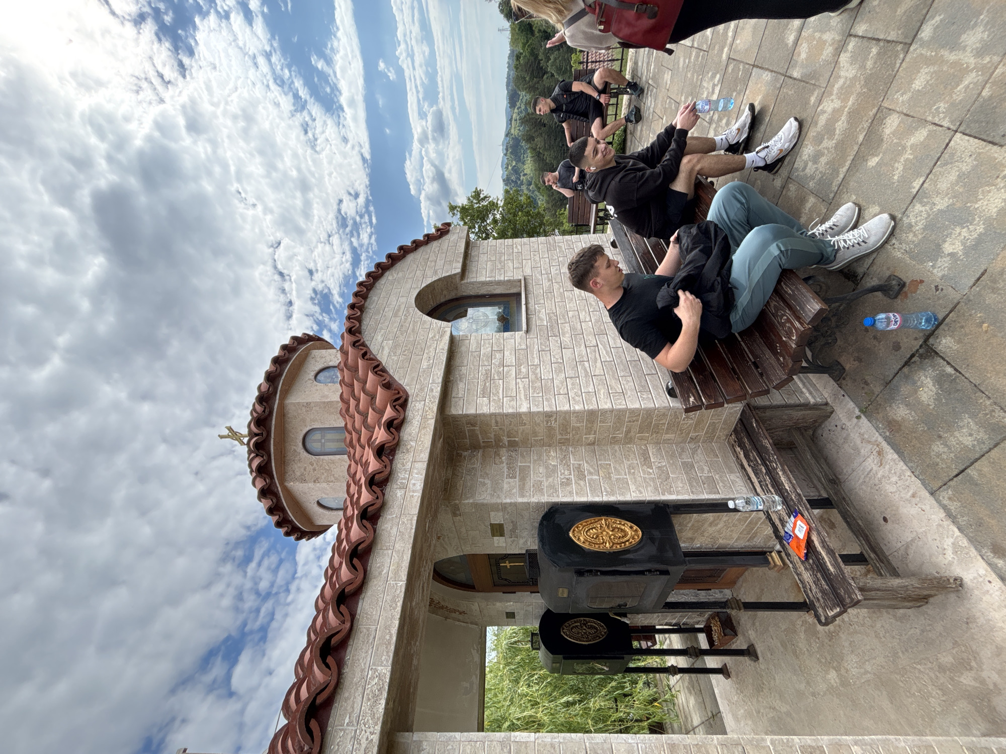 Outside a stone structure with a terracotta-tiled roof, in Zlatograd, Bulgaria, four young men are sitting on benches. The first man is closest, wearing a black t-shirt and blue-green pants. Another man beside him is wearing a black hoodie and beige shorts. The next man on the bench behind him is wearing a black t-shirt and shorts, with white sneakers. Further behind, another man is similarly dressed, sitting at the edge of the benches. One has a bottle of water. A church with a golden cross is visible to the left and behind the men. The sky is partly cloudy. There are wooden benches and donation boxes outside the building.
