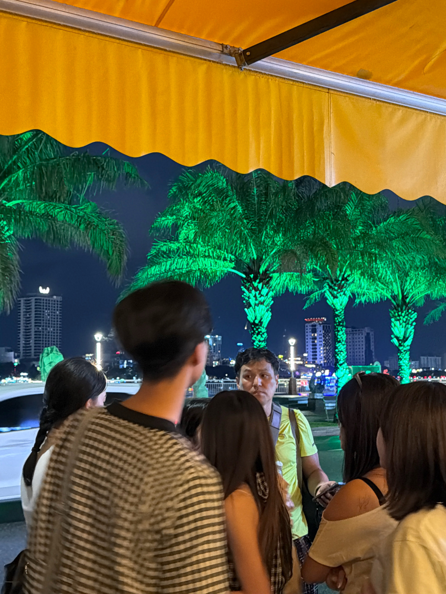A group of people are gathered outdoors under an orange awning at night. Palm trees illuminated with green lights are visible in the background, along with city buildings. A man in a lime green shirt stands behind several younger individuals. The scene appears to be in the city of Đà Nẵng, Vietnam, likely along the Han River, given the presence of illuminated palm trees and the cityscape characteristic of that area.