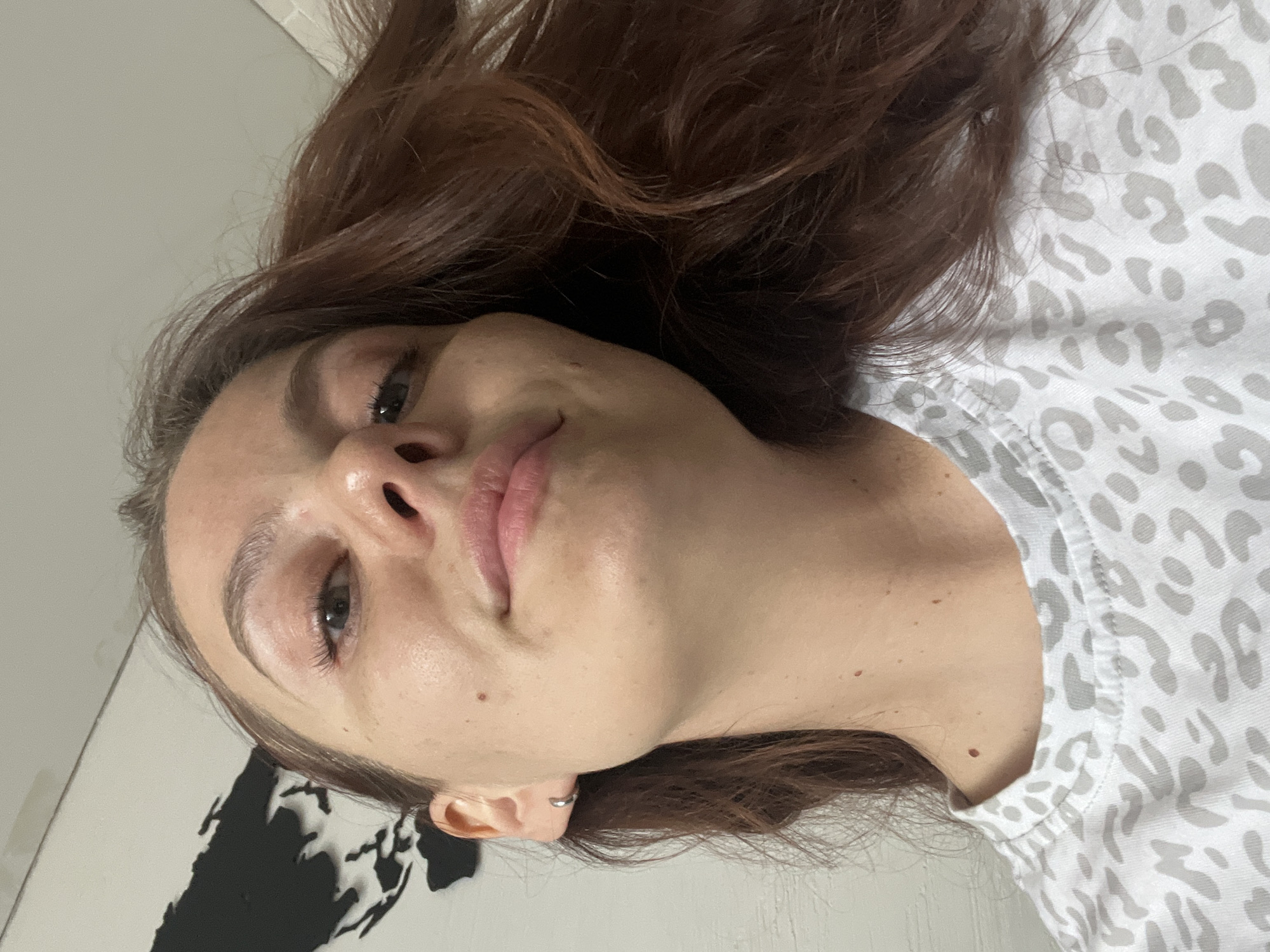 A close-up, bottom-up portrait captures a young woman lying down and looking directly at the camera. Her head is tilted, with her dark brown hair spread out around her. She has light skin, full pink lips, and visible eyelashes, with a small silver hoop earring in her right ear. She is wearing a light-colored t-shirt adorned with a grey leopard print pattern. Her expression is subtle, appearing relaxed and content. The background consists of a plain, light-colored surface, possibly a bed or floor. To her left, a black and white stylized outline, resembling a map or a landmass, is partially visible. The lighting is soft and even, indicating an indoor setting. There are no specific cues for time of day or weather. No visible text is present in the image. The location context for this image is Chaikovsky, Russia.