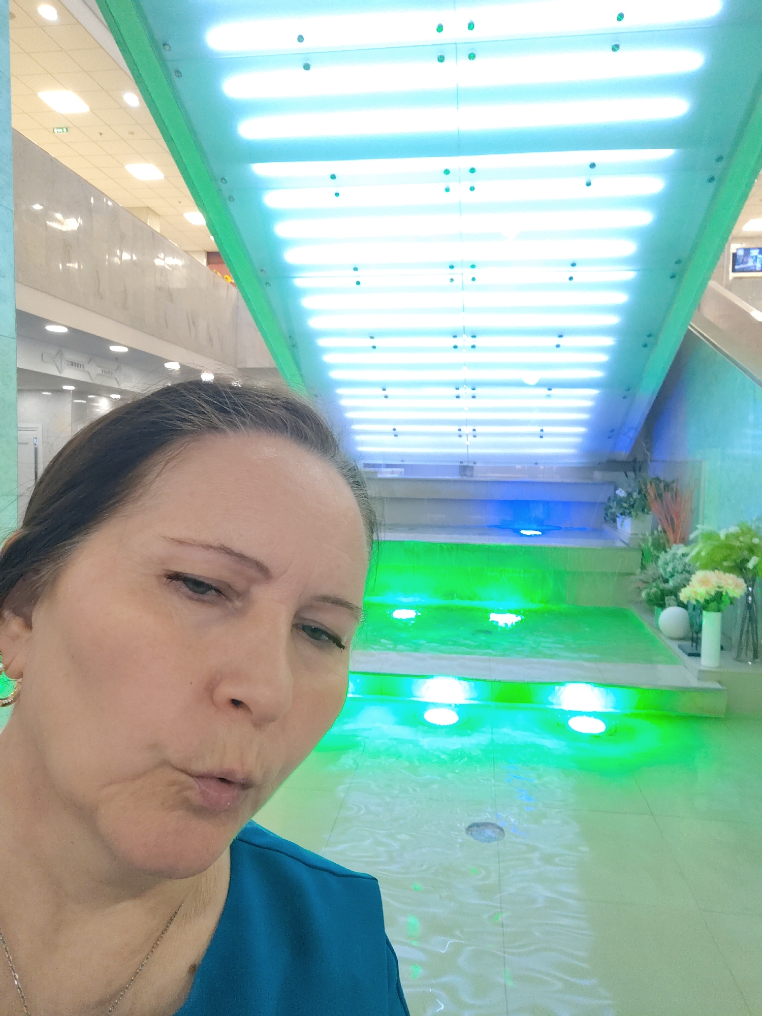 A woman is captured in a selfie-style shot, her face filling the left side of the frame, with an indoor public space in the background. She appears to be middle-aged, with her dark hair pulled back. Her left ear is visible, adorned with a gold hoop earring, and a thin silver chain necklace hangs around her neck. She wears a teal-colored top. Her mouth is slightly open, as if she is about to speak or making a puckered expression, and her gaze is directed slightly downwards and to the right.
The background features a vibrant and modern architectural design. A multi-tiered water feature or decorative fountain dominates the center-right, illuminated by bright green and blue lights. The lower pool of water glows a vivid green, with reflections of unseen light sources, while the upper cascade appears blue. Above this, an angled ceiling structure extends upwards and to the right, adorned with numerous bright, horizontal white or light-blue fluorescent tubes or LED panels. Small circular dots, possibly fasteners or additional small lights, are visible on this structure.
The broader setting suggests a well-lit, contemporary indoor environment, possibly a shopping mall or a large commercial building, consistent with a modern space in Moscow. In the further background, a light-colored tiled ceiling with recessed lighting is visible, along with walls that appear to be paneled or made of light stone. A staircase can be glimpsed on the far right. Decorative white and green artificial plants are arranged around the water feature. The lighting is entirely artificial, creating a striking and luminous atmosphere. No legible text is visible in the image.