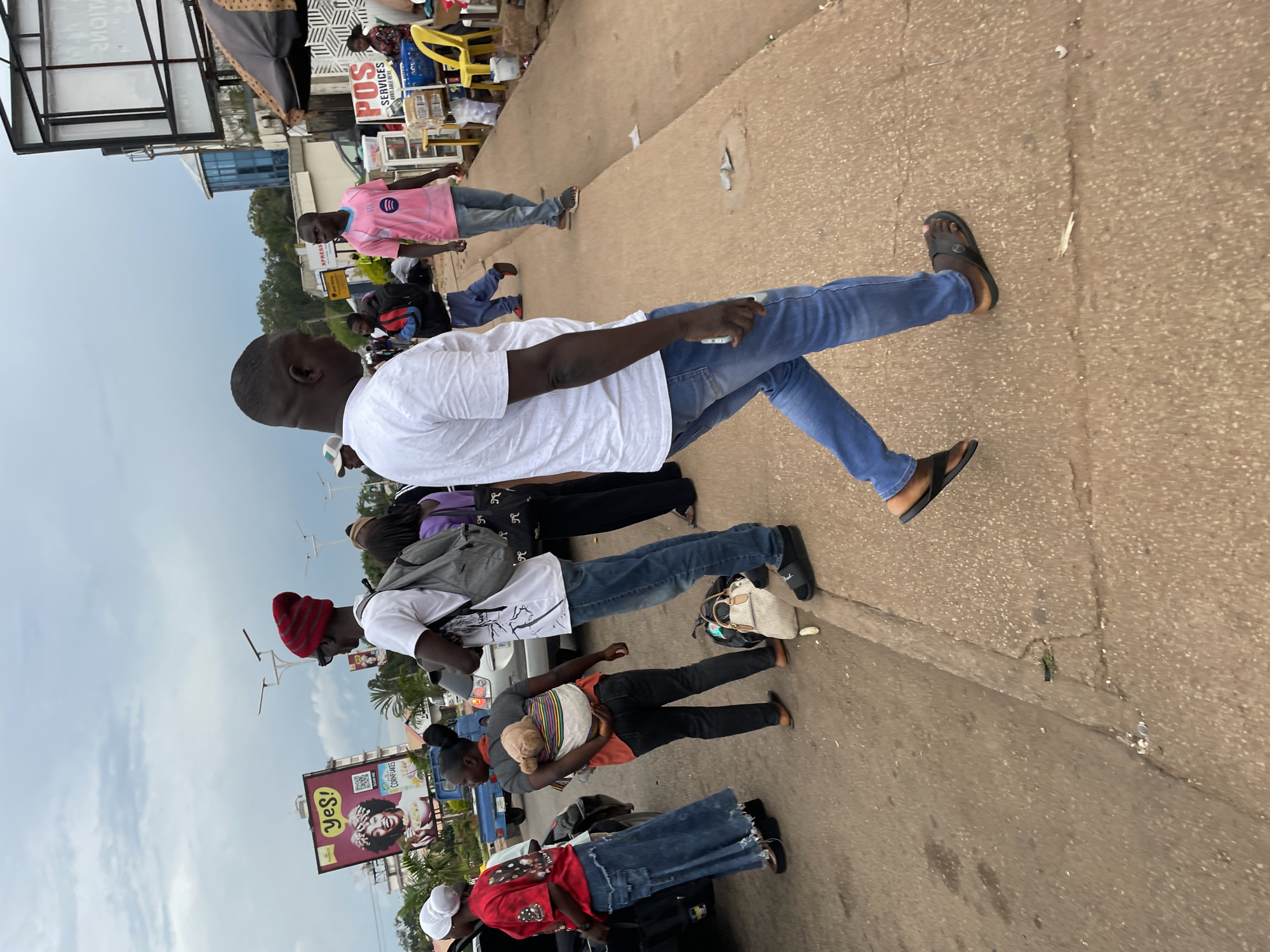 In Jos, Nigeria, on a cloudy day, a diverse group of people are gathered on a paved sidewalk next to a road. The scene appears to be a busy commercial area, with a prominent billboard advertising "Yes!" and "Cornflakes" visible in the background.
In the foreground, a man in a white t-shirt and blue jeans walks away from the camera. He is wearing dark sandals. To his left, a woman in a red t-shirt and blue denim skirt, wearing a white headscarf, is standing near a car. Another woman, wearing a grey shirt and dark pants, is carrying a baby on her back, wrapped in a colorful cloth.
Further in the background, a man in a pink t-shirt and jeans is walking on the sidewalk. Other individuals, some carrying bags, are also present. Stalls with umbrellas and goods are set up along the sidewalk, suggesting a marketplace. The environment is characterized by a paved road and sidewalk, with some debris on the ground. The overall atmosphere is bustling and indicative of daily life in a Nigerian city.