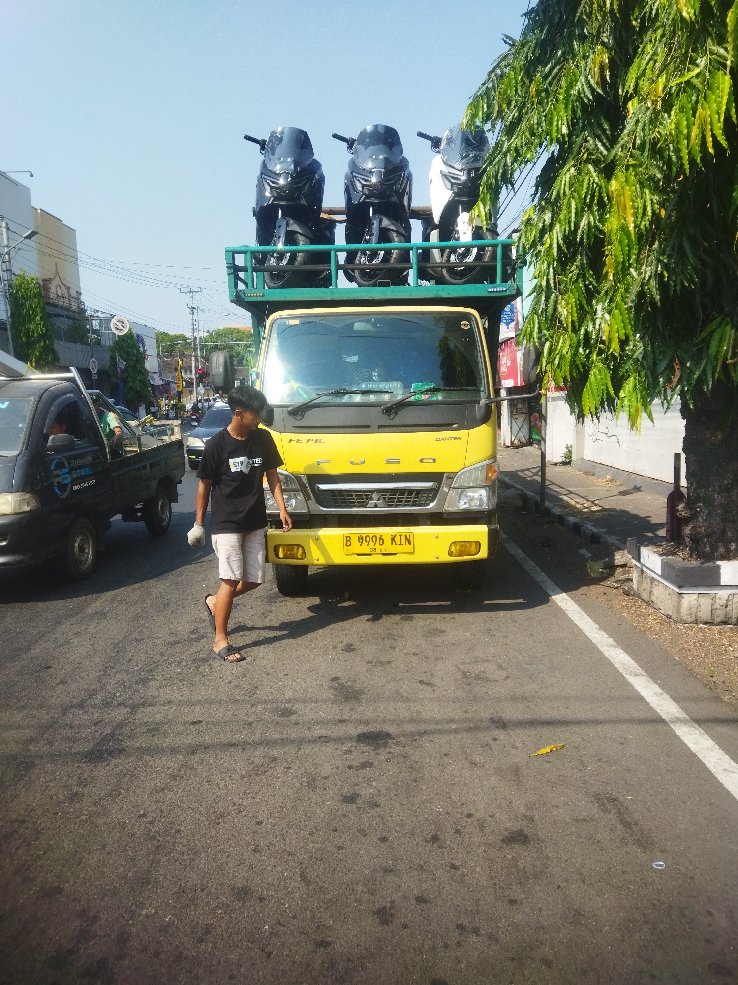 On a sunny day, on a street in Giripurwo, Indonesia, a yellow truck is parked. On the truck's flatbed, three black and white scooters are stacked vertically, secured by a green metal railing. In front of the truck, a young man in a black t-shirt and light-colored shorts walks from right to left. He is wearing white gloves on his right hand and flip-flops on his feet. To the left of the yellow truck, a black pickup truck is also parked. The street is paved and lined with a white stripe on the right side. A large tree with green leaves stands to the right of the yellow truck, casting shadows on the ground. Buildings and power lines are visible in the background, indicating an urban or semi-urban environment.