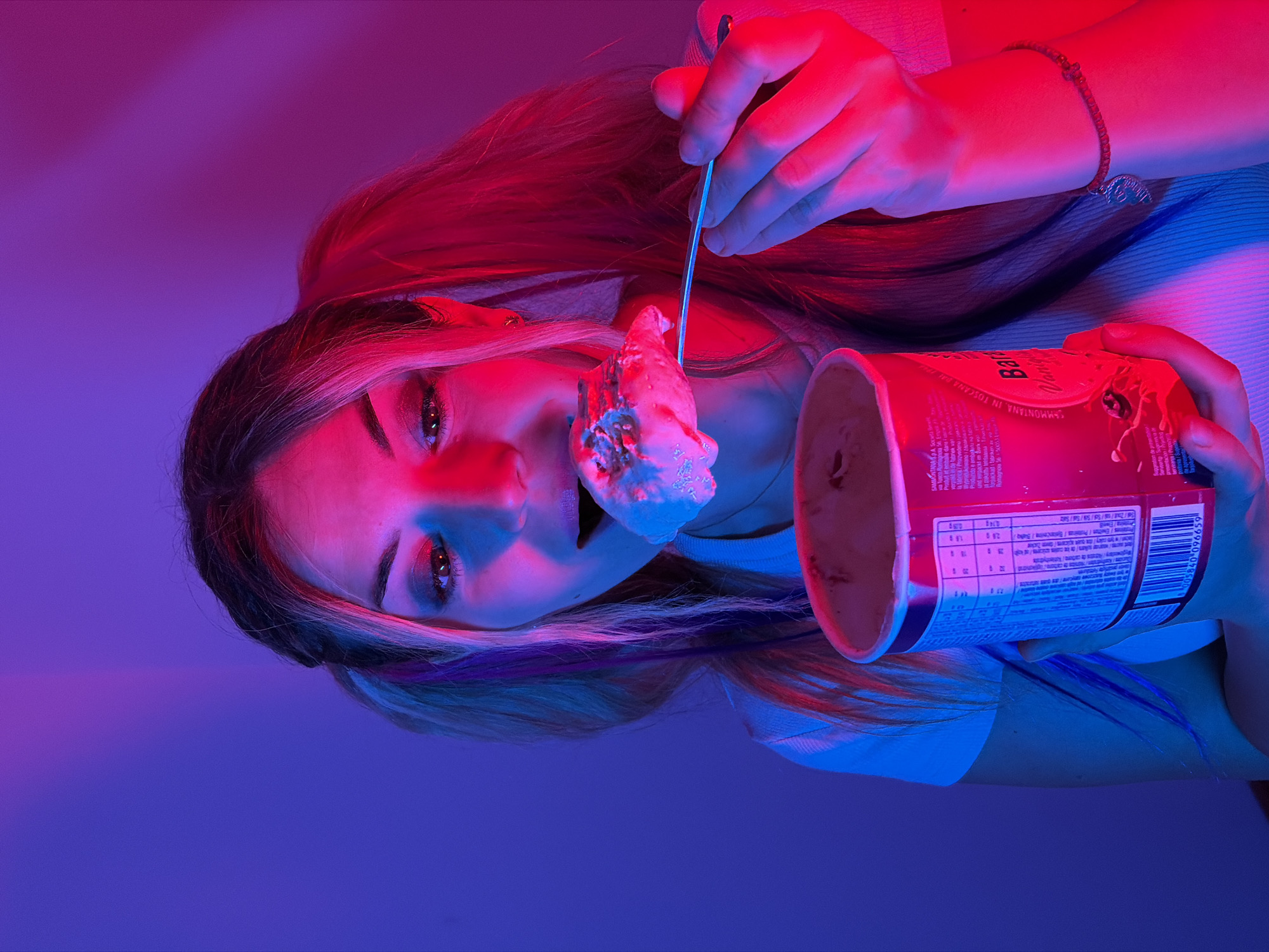 A young woman with blonde hair, styled in two pigtails with streaks of purple and pink, is eating ice cream in a dimly lit room. She is holding a red tub of ice cream in her left hand and a spoon with a scoop of ice cream in her right hand, bringing it to her mouth. She is wearing a white top. The room is illuminated with purple and red lighting, creating a vibrant and moody atmosphere. The background is a plain wall. No specific location in Budva, Montenegro is identifiable.