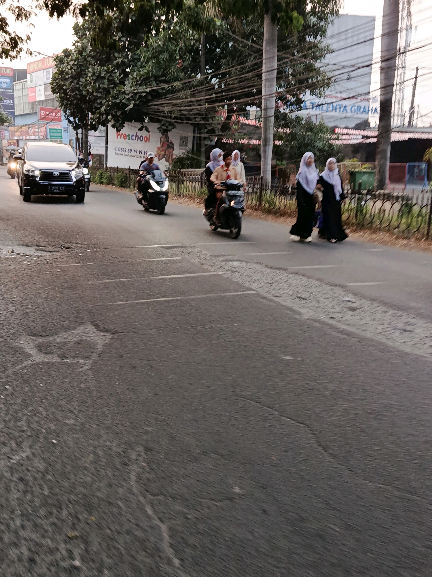 The image captures a street scene in Bekasi, Indonesia, likely during daytime given the lighting. In the foreground, the asphalt road shows signs of wear, with visible potholes and cracks. Several vehicles are present, including a dark-colored SUV on the left side of the road and multiple motorcycles. A black SUV with license plate B 2895 KZH is prominent, moving forward. On the right side of the road, three women in hijabs, dressed in black, are walking. One is partially obscured by the other. Further down the road, a motorcycle with two individuals on it is moving away from the camera, and another motorcycle with a driver is also visible. In the background, there are buildings, trees, and overhead power lines. A banner for "Preschool" with a phone number and "Creative Curriculum (USA)" is displayed on a billboard. Another building in the distance has "TALENTA GRAHA" written on it. The overall environment suggests a busy urban or suburban area.
