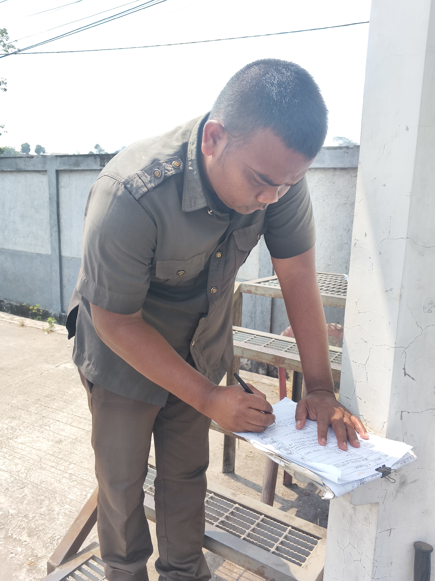A man wearing a dark olive short-sleeved uniform shirt and dark brown pants is standing and writing on a stack of papers. He is holding a black pen in his right hand and his left hand is resting on the papers. The papers are on a light-colored clipboard, which is resting on a metal grate platform. The man has short dark hair and is looking down at the papers. The background shows a concrete wall, some vegetation, and a bright sky with some overhead wires. The scene appears to be outdoors, possibly in a public or industrial area.