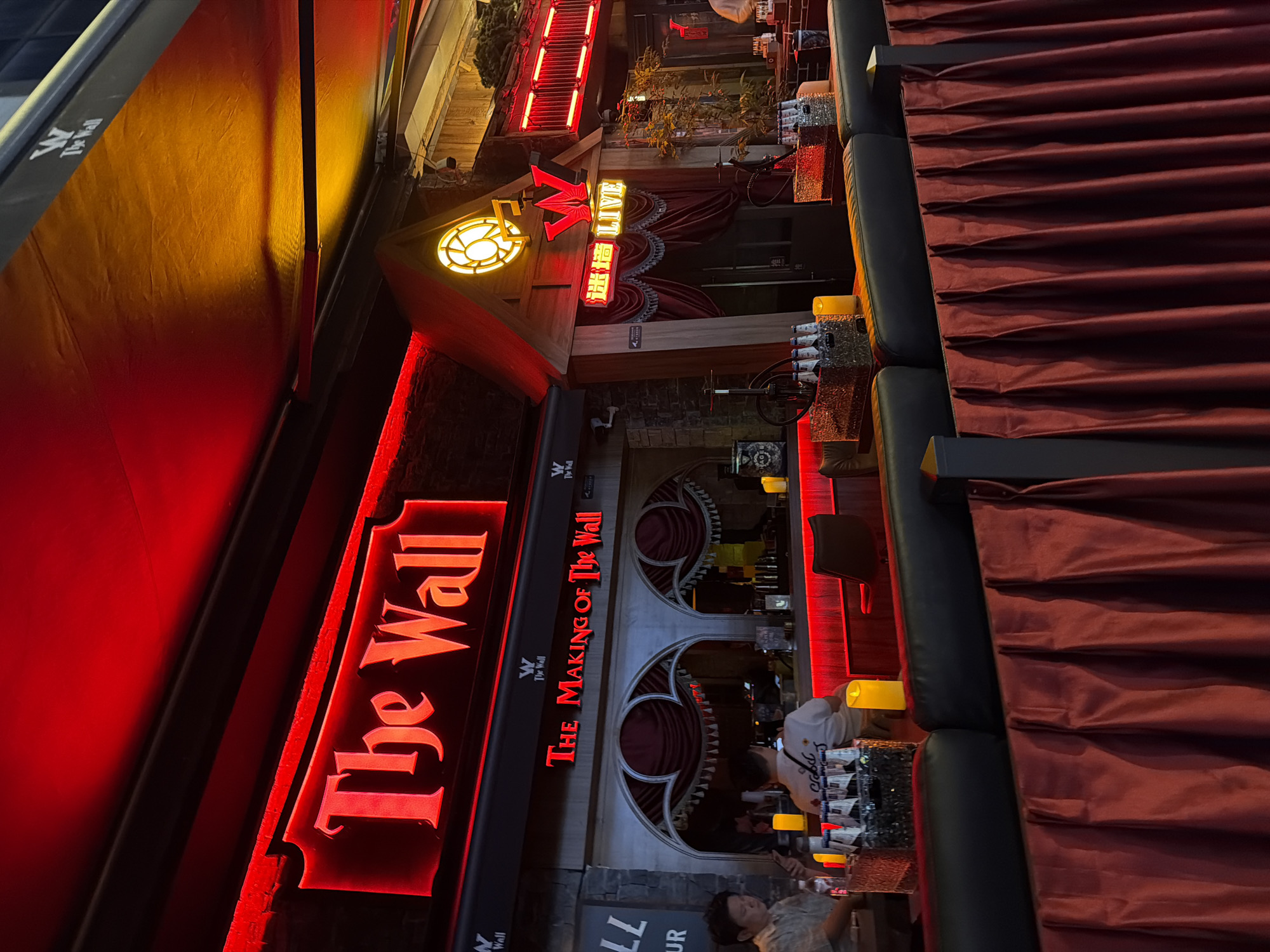 Here's a description of the image:
The image captures the facade of "The Wall" bar in Liangjiang, China. The bar's name is prominently displayed in red neon lettering, stacked vertically. The sign also reads "THE MAKING OF The Wall". The entrance area is decorated with architectural elements suggestive of arches and drapes. The scene is illuminated with a mix of red neon lights and warmer, candle-like lights on the bar. A few people are visible inside the bar. Some are standing near the bar area, seemingly engaging with bartenders.
Outside the bar, a retractable awning is partially extended, casting a warm yellow glow. To the side of the awning there are curtains draped. The image is shot from a somewhat oblique angle, with the camera tilted.