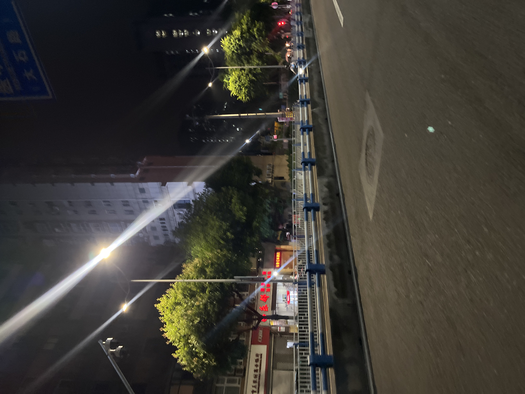 This is a night-time, eye-level, medium shot of a street in Jiulongpo District, China. The image is characterized by strong light streaks from streetlights and building lights, indicating a long exposure or motion blur. On the left side of the image, a tree with vibrant green foliage is illuminated, with a street light and what appears to be a security camera pole also visible. Behind the tree, a multi-story building with numerous windows is dimly lit. A street sign with blue and white Chinese characters is visible in the upper right corner. The foreground features a roadway with faint lane markings and a concrete barrier with blue and white railings separating the road from a sidewalk or elevated area. Several businesses with illuminated signs are visible behind the barrier. The overall atmosphere is urban and nocturnal, with a focus on the contrast between light and shadow.