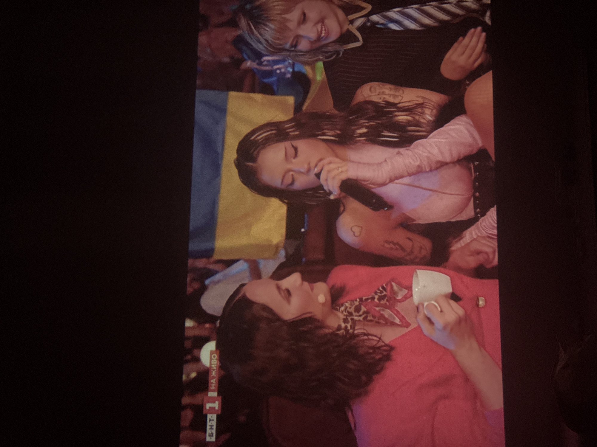 Here is a description of the image:
The image shows three women in what appears to be a television broadcast setting. In the bottom of the image is a woman with dark curly hair, wearing a red outfit with a patterned scarf, and holding a white cup. In the center is a woman with long, dark, wavy hair holding a microphone and singing. She is wearing a light pink outfit. Behind her is a Ukrainian flag. At the top of the image is a woman with blond hair, wearing a striped top, and smiling. The broadcast appears to be live, based on the text visible on the screen. The location is unknown.