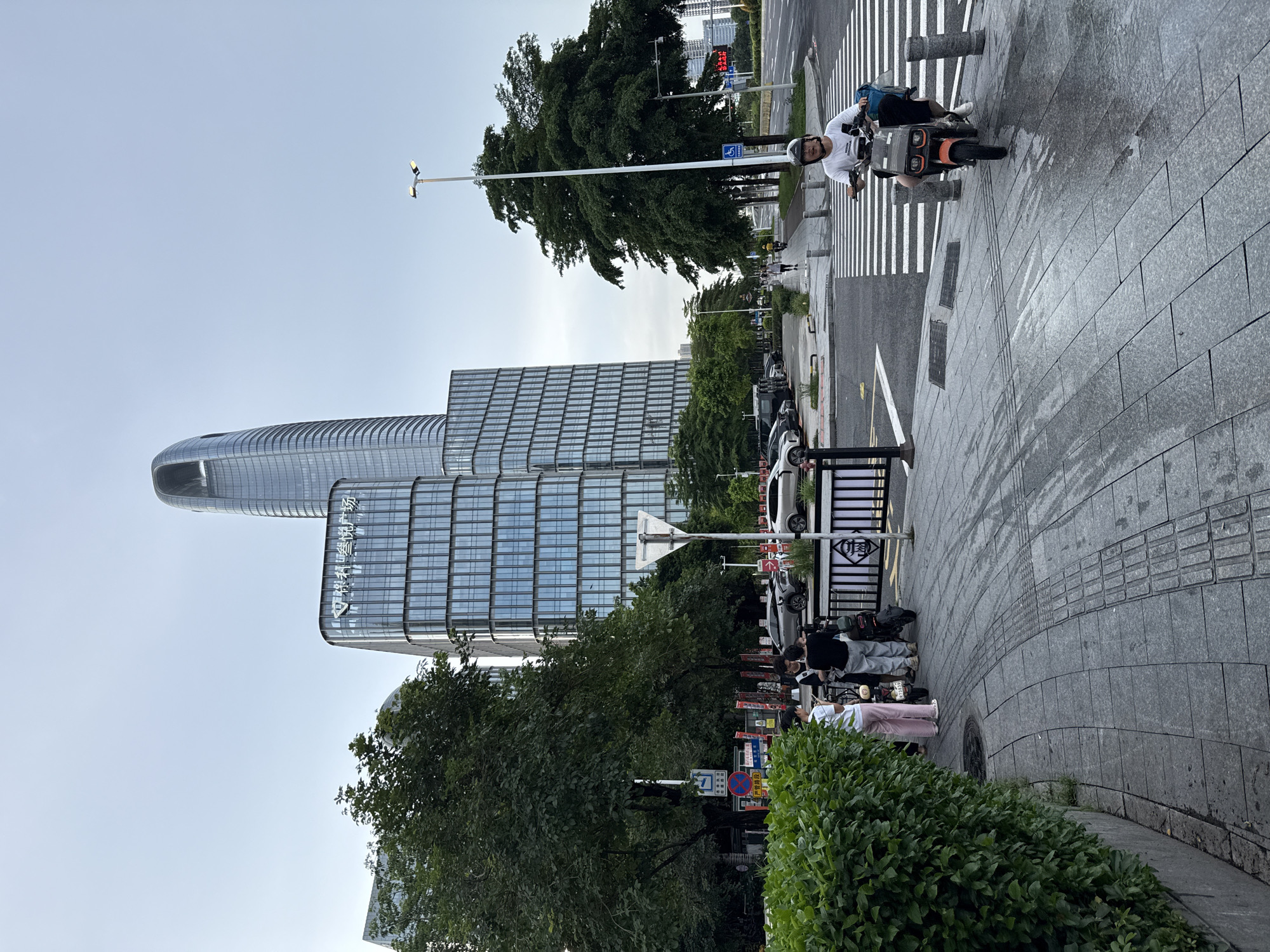 In Haizhu District, China, on a cloudy day, a street scene unfolds with modern architecture and daily life. Dominating the skyline are several contemporary glass-paneled skyscrapers, including a distinctive curved tower with an open top. In the foreground, a pedestrian walkway is paved with grey tiles, some of which are textured for tactile paving.
To the left, a dense green hedge borders the walkway. A group of three individuals are gathered near a parked electric scooter and a motorcycle, engaged in conversation. One person is wearing a white and pink striped shirt and pink pants, another a black shirt and grey pants, and the third, a black shirt and black pants.
Further down the street, a man wearing a white t-shirt, black shorts, and a helmet rides an orange electric scooter across a pedestrian crossing. He has a blue bag on the back of the scooter. Cars are parked along the roadside, and trees line the street, providing shade. Streetlights are visible, and traffic signals indicate the presence of a road intersection. The overall impression is one of an urban environment where modern development coexists with everyday transportation and pedestrian activity.