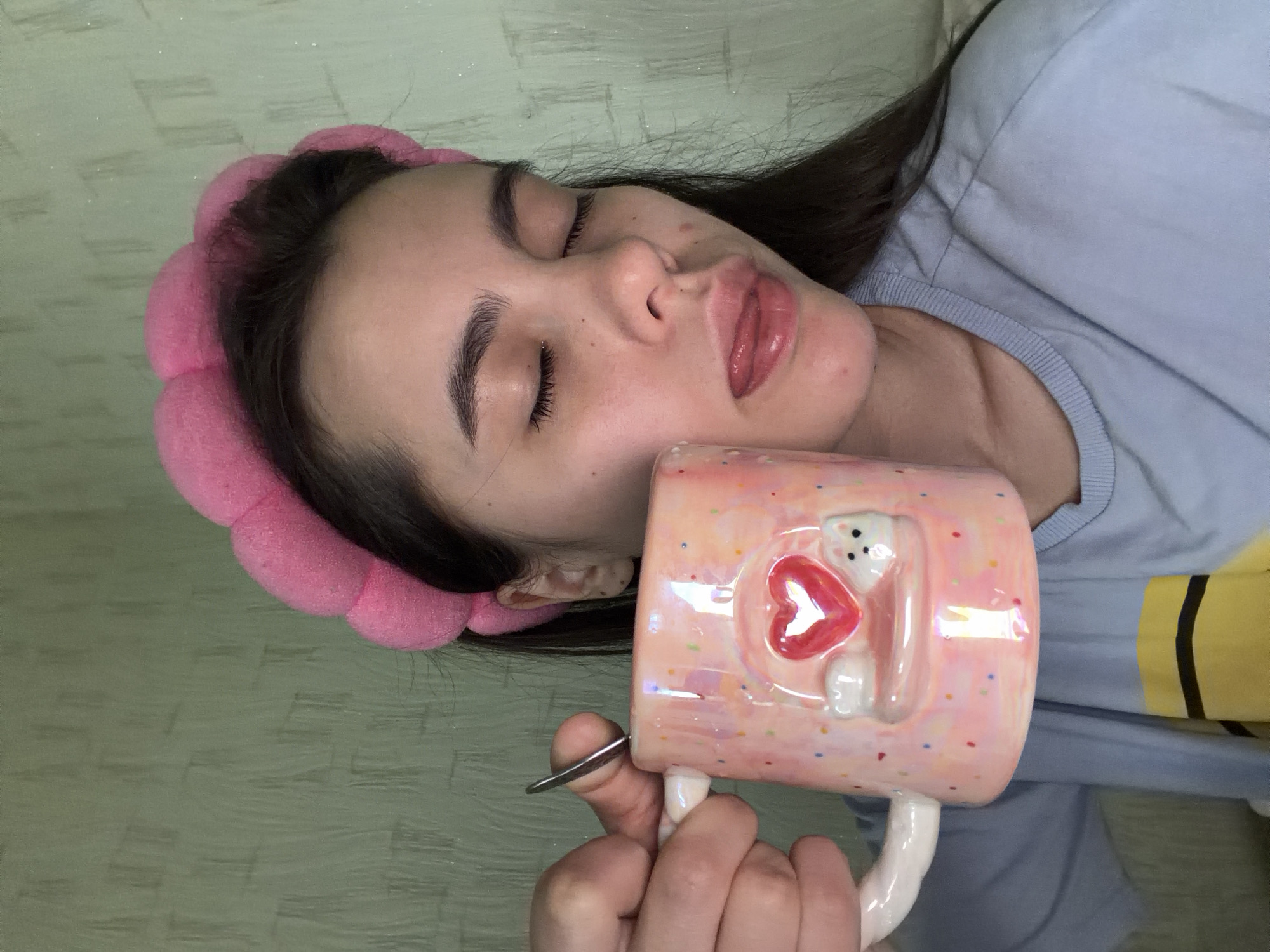 An indoor scene in Chaikovsky, Russia, features a young woman with dark hair, eyes closed, and tongue slightly extended, wearing a bright pink padded headband and a light blue t-shirt with a yellow and black graphic. She holds a glossy, iridescent pink ceramic mug adorned with colorful speckles, a prominent red heart, and a small white animal figure in her left hand. A silver spoon is partially visible within the mug. The background consists of a textured, light green wall.