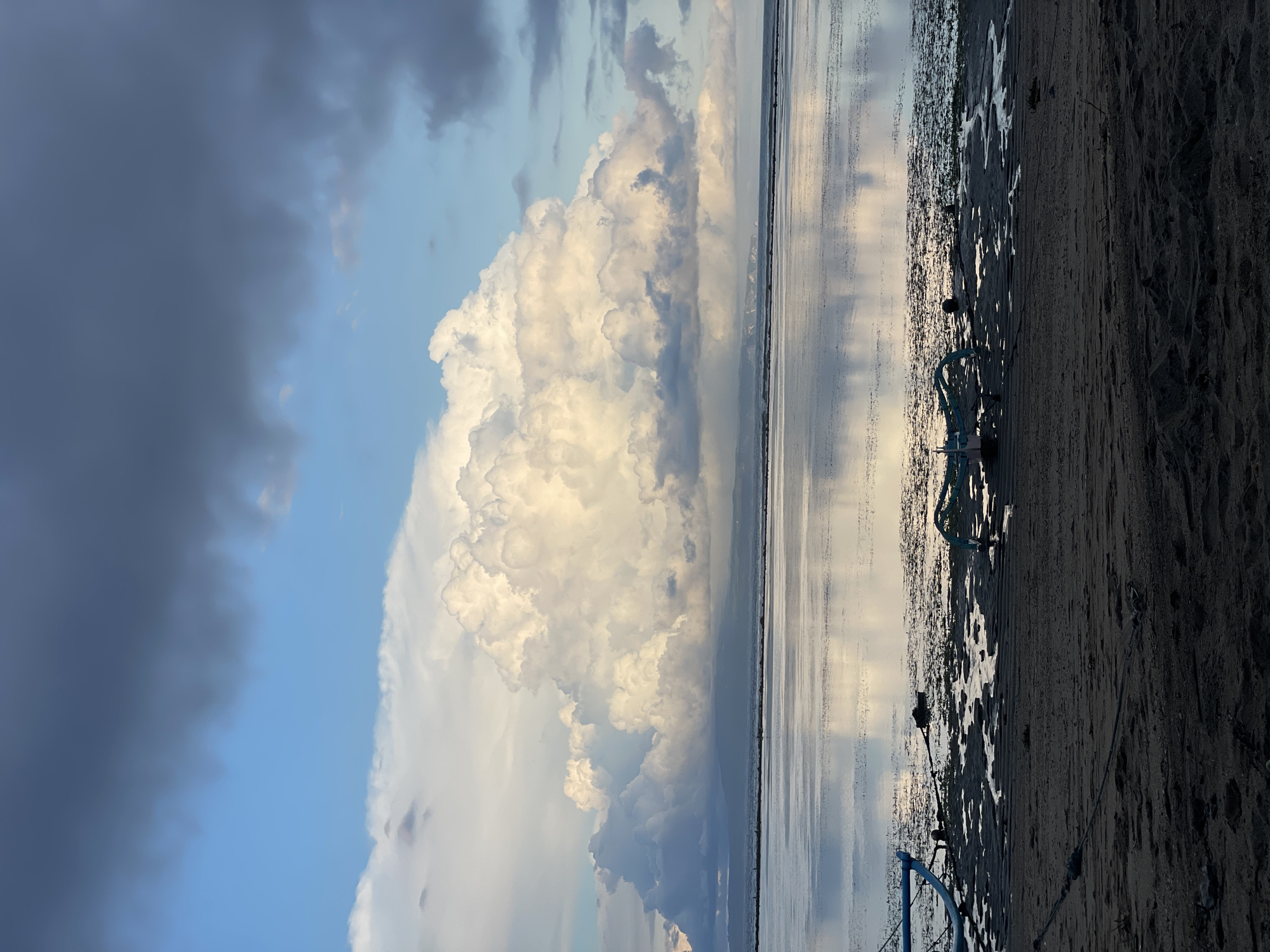 The photograph captures a beach landscape, likely at low tide, with a prominent cloud formation dominating the sky. The left side of the frame reveals a blue sky partially obscured by dark gray clouds. A massive cumulus cloud formation, illuminated by sunlight, stands out against the lighter sky, mirrored in the shallow water covering the flat beach. The water surface reflects the sky and clouds, creating a sense of depth and vastness. The beach itself is a mix of sand and exposed mud flats, with traces of water creating a mottled texture. In the foreground, a portion of a blue object (possibly a structure or equipment) is visible at the bottom left, along with some indistinct objects along the shoreline. The scene appears deserted, with no people visible. The lighting suggests either sunrise or sunset. Given the location in Denpasar, Indonesia, and the low-lying coastal environment, it's likely a scene from one of the beaches in the vicinity, potentially affected by tidal fluctuations.