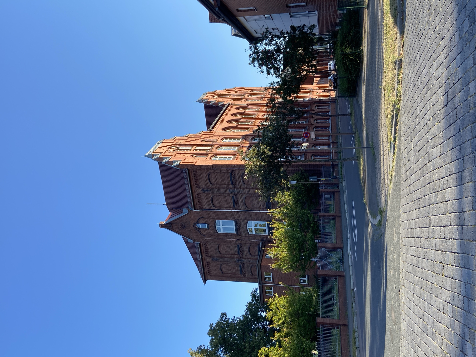 The image depicts a brick building with a red tiled roof, set against a clear blue sky. The building has a traditional architectural style with numerous arched windows and decorative brickwork. In front of the building, there is a metal fence and a row of trees with green foliage. A paved road and sidewalk are visible in the foreground, with a few parked cars in the background. The scene appears to be a residential or institutional area in a city, under the bright illumination of daylight.