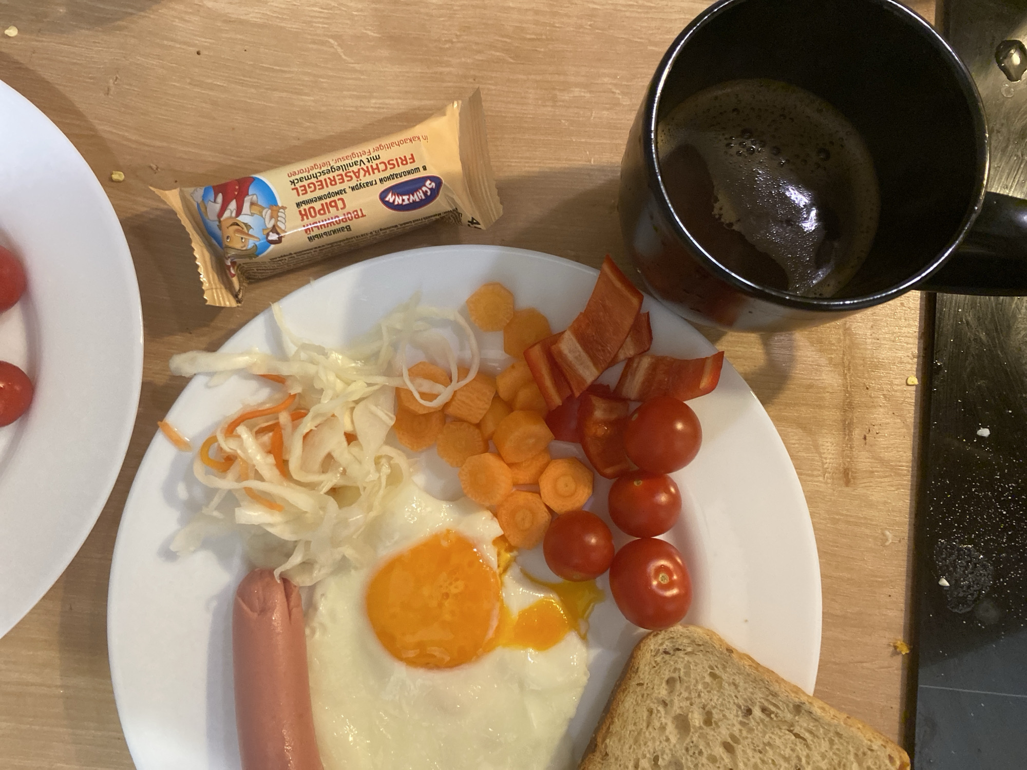 The image shows a breakfast meal on a white plate, consisting of a fried egg, sausage, toast, sliced vegetables (carrots, red peppers, tomatoes, cabbage), and a vanilla-flavored chocolate bar. A black mug filled with coffee is also present. The breakfast is set on a wooden surface. A portion of another white plate is visible on the left. The presence of the "Schwinn" chocolate bar with text in both Russian and German suggests the breakfast setting might be located in a region where both languages are common or where products from those regions are readily available. Based on the available information, it's impossible to determine the exact location in Budva, Montenegro, or even confirm that it is in Budva.