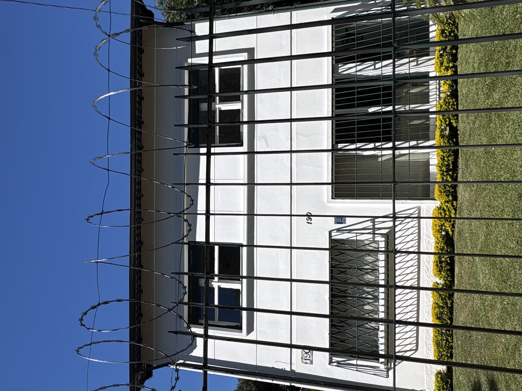 A white, two-story building is visible behind a metal fence with razor wire on top. The building has several windows, some with metal bars. There are two house numbers visible, "190" on the left side of the building and "190" on the right. A row of yellow bushes runs along the base of the building, and a green lawn is in front of the fence. The sky is clear and blue. The scene appears to be in the city of São Paulo, Brazil, given the architectural style and context.