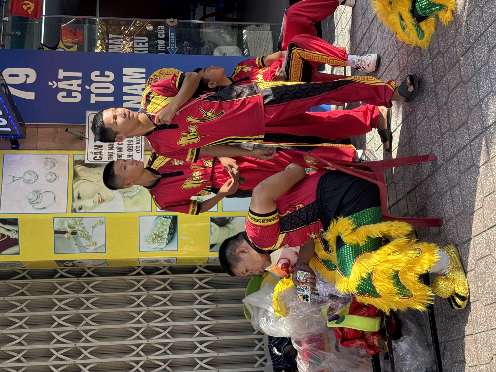 The image depicts a street scene in Nha Trang Ward, Vietnam, on a sunny day. In the foreground, a group of young men, likely performers, are dressed in vibrant red and yellow costumes. One man in the center is holding a smartphone and looking at it. Another man to his left is seated and looking away, partially obscured by a large, fluffy yellow and green costume piece that resembles a lion's head. Several other individuals in similar red and yellow attire are visible, some seated and others standing. Behind them, a yellow pillar displays images of jewelry, including necklaces and earrings, suggesting a nearby jewelry store. A metal roll-up gate is visible to the left, and a blue sign with white lettering that reads "CẮT TÓC NAM" (Men's Haircut) is present on the right. The ground is paved with interlocking stones. The overall atmosphere suggests a pause or preparation period for a performance or event.