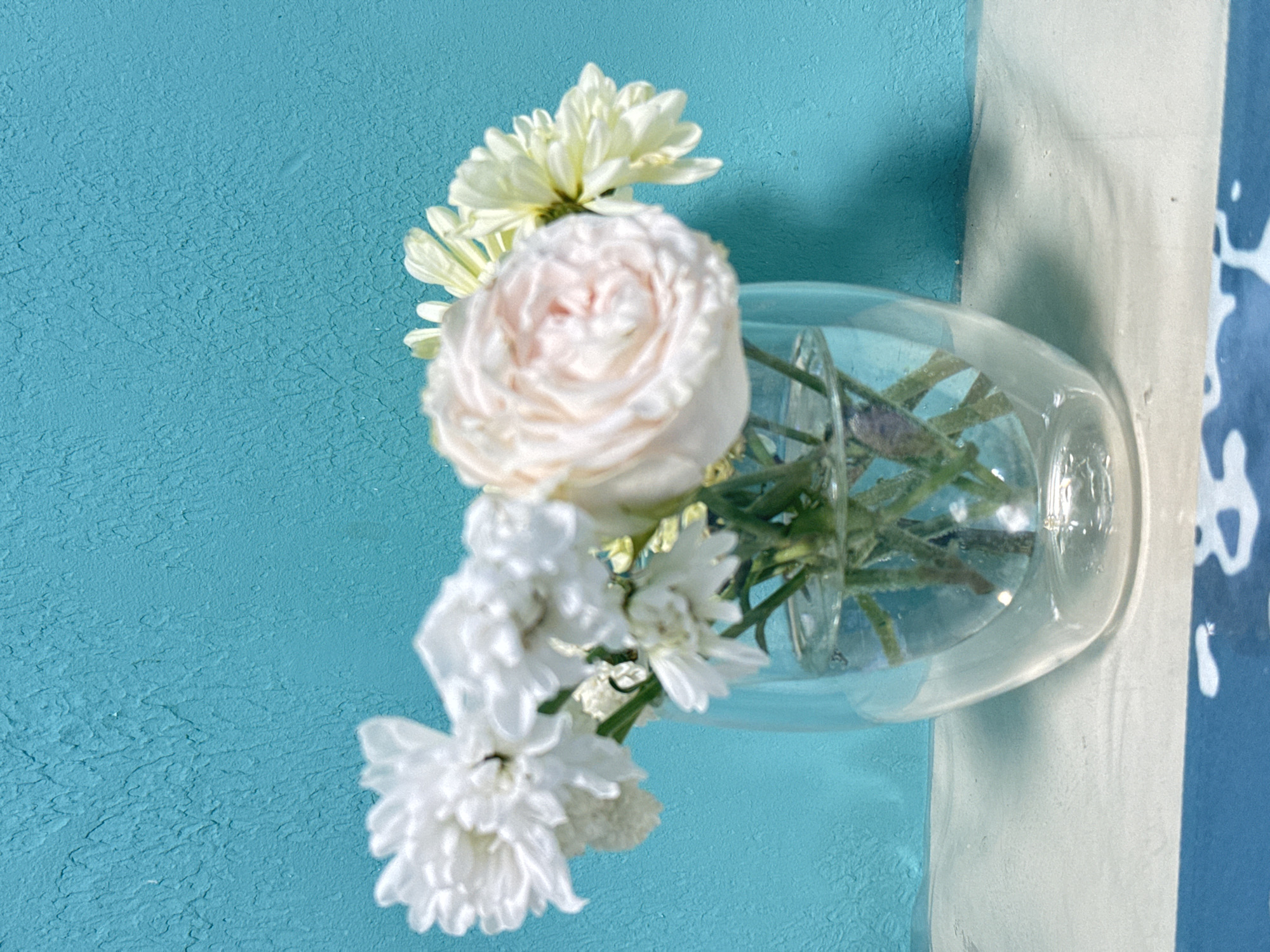 A small, clear glass vase containing a floral arrangement is positioned against a turquoise wall. The arrangement features a pale pink rose, a white chrysanthemum, and small white flowers. The vase rests on a white surface next to a blue and white patterned object.
There is no information in the image to identify the location as Zav'yalovo, Russia.