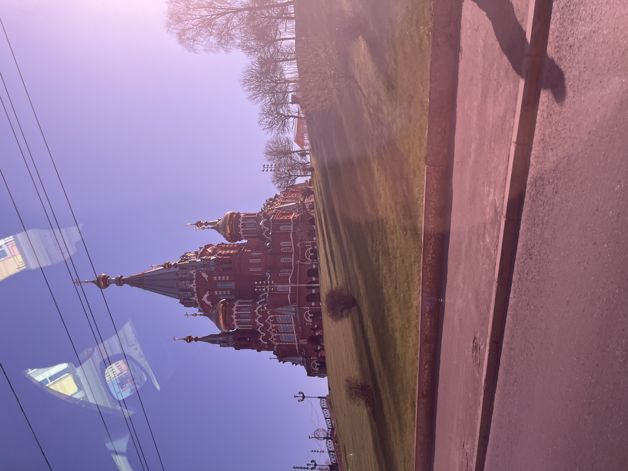 The image shows the Cathedral of Archangel Michael in Izhevsk, Russia. The red-brick cathedral features multiple domes and ornate detailing. A grassy area slopes towards the building, with trees visible in the background. Power lines are present in the foreground. A portion of a vehicle's window and the shadow of a person are also visible.