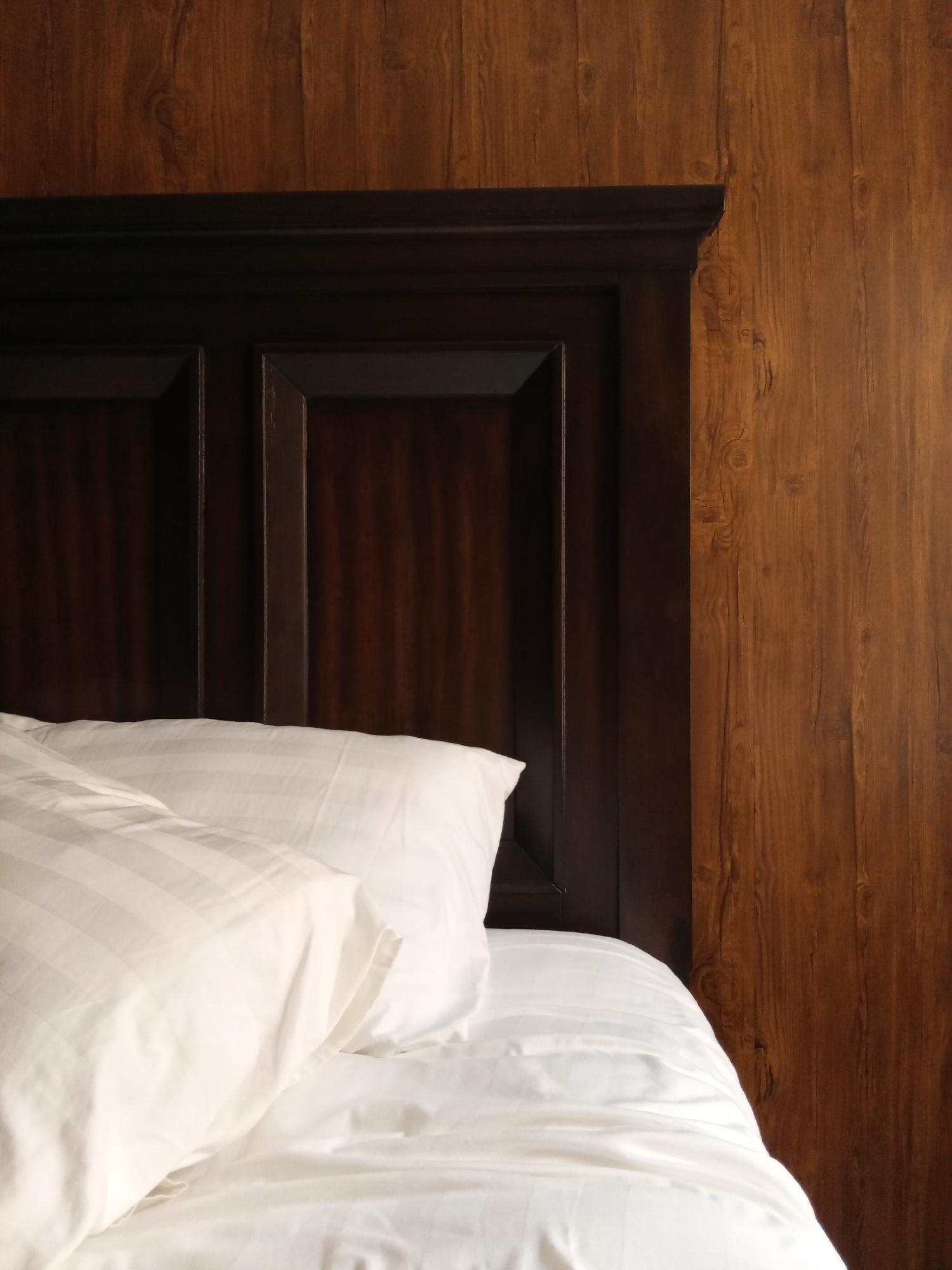The image depicts an interior scene, likely a hotel room in Nha Trang, Vietnam. The foreground is dominated by white bedding, including two white pillows with subtle stripe patterns and a white duvet or sheet, appearing slightly disheveled. Behind the bedding, a dark brown, ornate wooden headboard is visible, featuring recessed rectangular panels and a molded top edge. To the right of the headboard and extending to the right edge of the frame, a vertical section of wood-paneled wall is present, characterized by a medium brown hue and prominent wood grain patterns. The lighting suggests ambient illumination, highlighting the texture of the bedding and wood. No people or specific actions are depicted. The precise location within Nha Trang is not identifiable from the visual information provided.