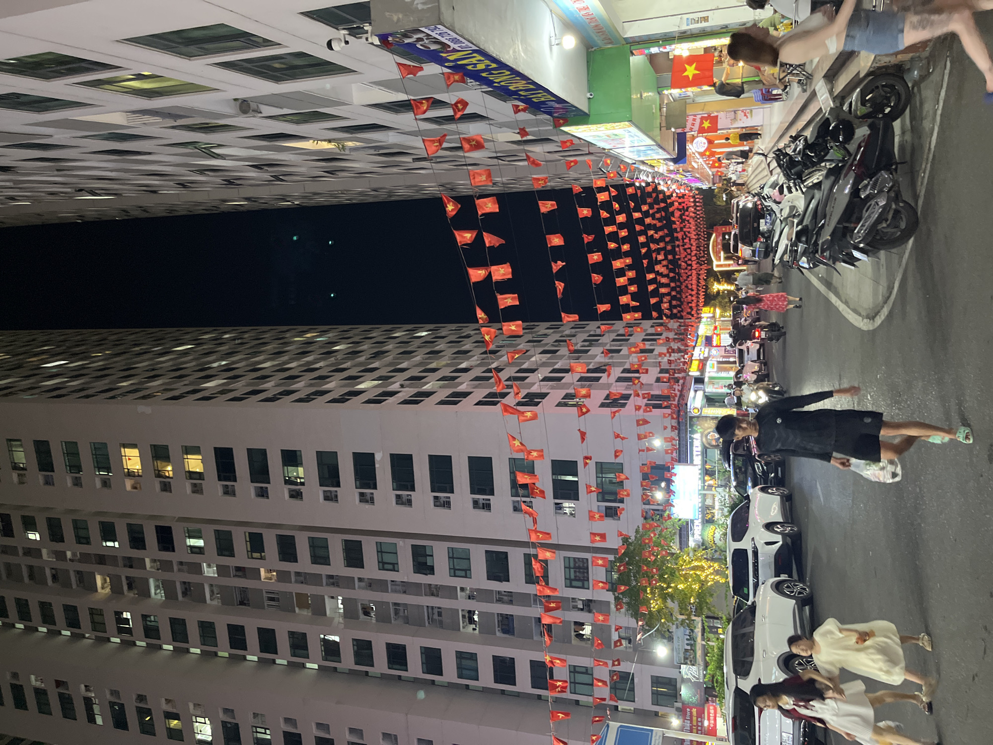 The image captures a street scene at night in Bac Nha Trang Ward, Vietnam. Two tall apartment buildings, one on the left and one on the right, flank a street lined with shops and businesses. Red flags with yellow stars, likely national flags, are strung across the street between the buildings, creating a festive atmosphere.
On the street, several vehicles are visible, including white SUVs and black cars, suggesting traffic. Motorbikes are parked on the side of the road and some are also in motion. Pedestrians are also present, walking on the sidewalk and crossing the street. On the left side of the street, two women and a young girl are walking together. A man in a black shirt and shorts is walking across the street in the foreground, carrying a plastic bag. On the right side, several people are seen near the shops.
The buildings have numerous windows, many of which are illuminated, indicating residential occupancy. The shops on the ground floor display signs and lights, suggesting commercial activity. A tree decorated with lights is visible between the buildings, adding to the nighttime ambiance. The sky is dark, typical of nighttime.